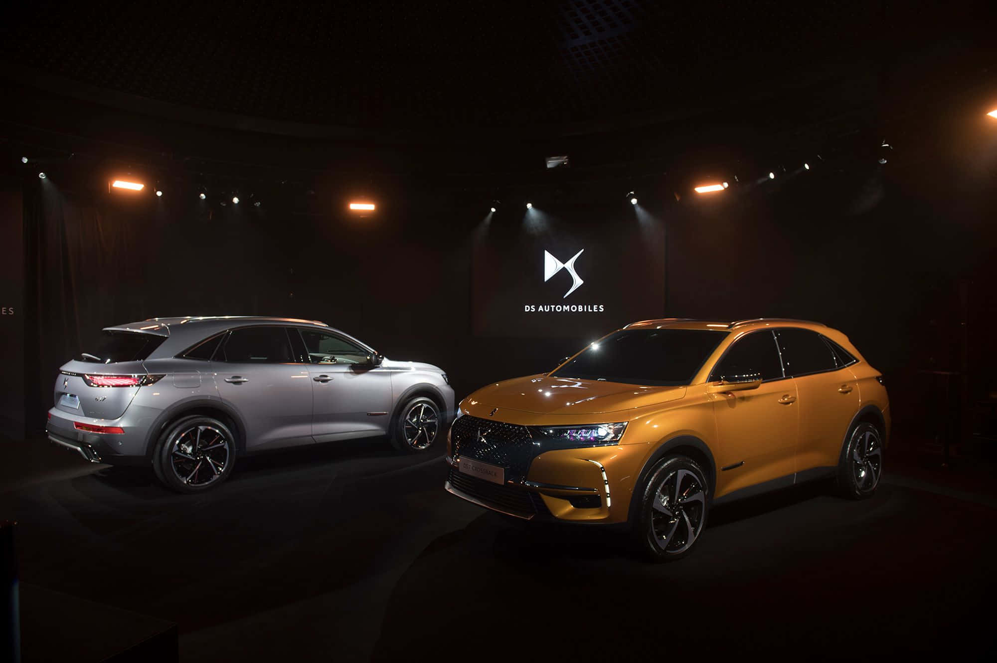 Ds Automobiles 桌布 壁紙 — 兩輛銀色和橙色的豪華SUV在黑暗舞台上戲劇性的聚光燈下閃耀，帶有DS Automobiles品牌標識。