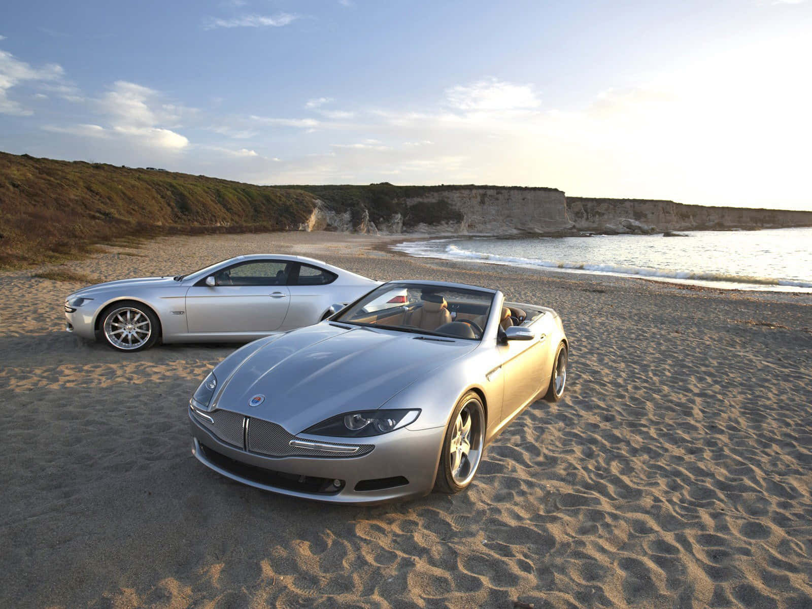 Fisker 桌布 壁紙 — 兩輛銀色豪華敞篷車停放在陽光明媚的沙灘上，背景有懸崖和海洋波浪。