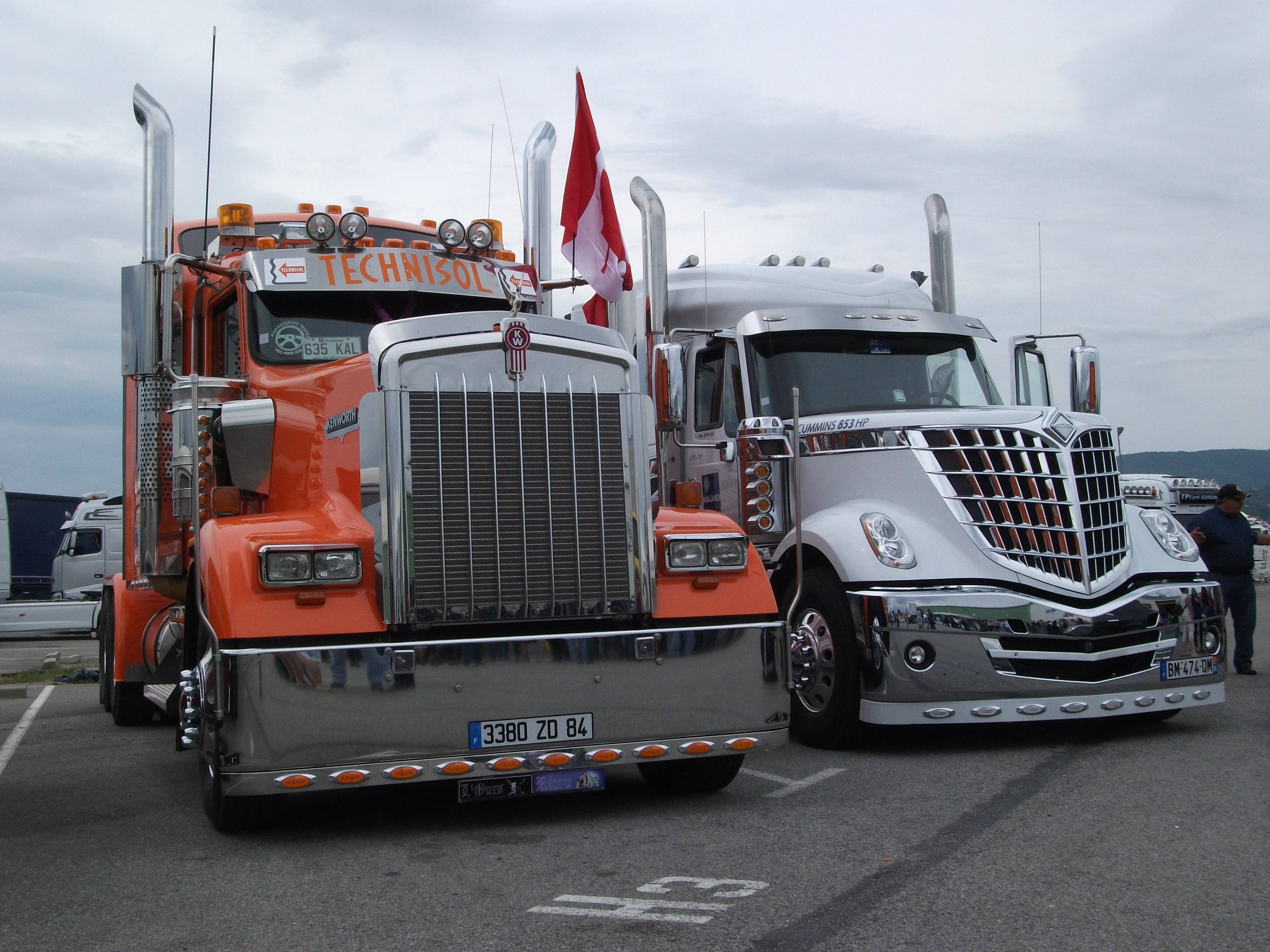Kenworth W900 桌布 壁紙 — 兩輛閃亮的半掛卡車，一輛橙色一輛銀色，並排停放在陰天天空下，加拿大國旗從橙色卡車上飄揚。