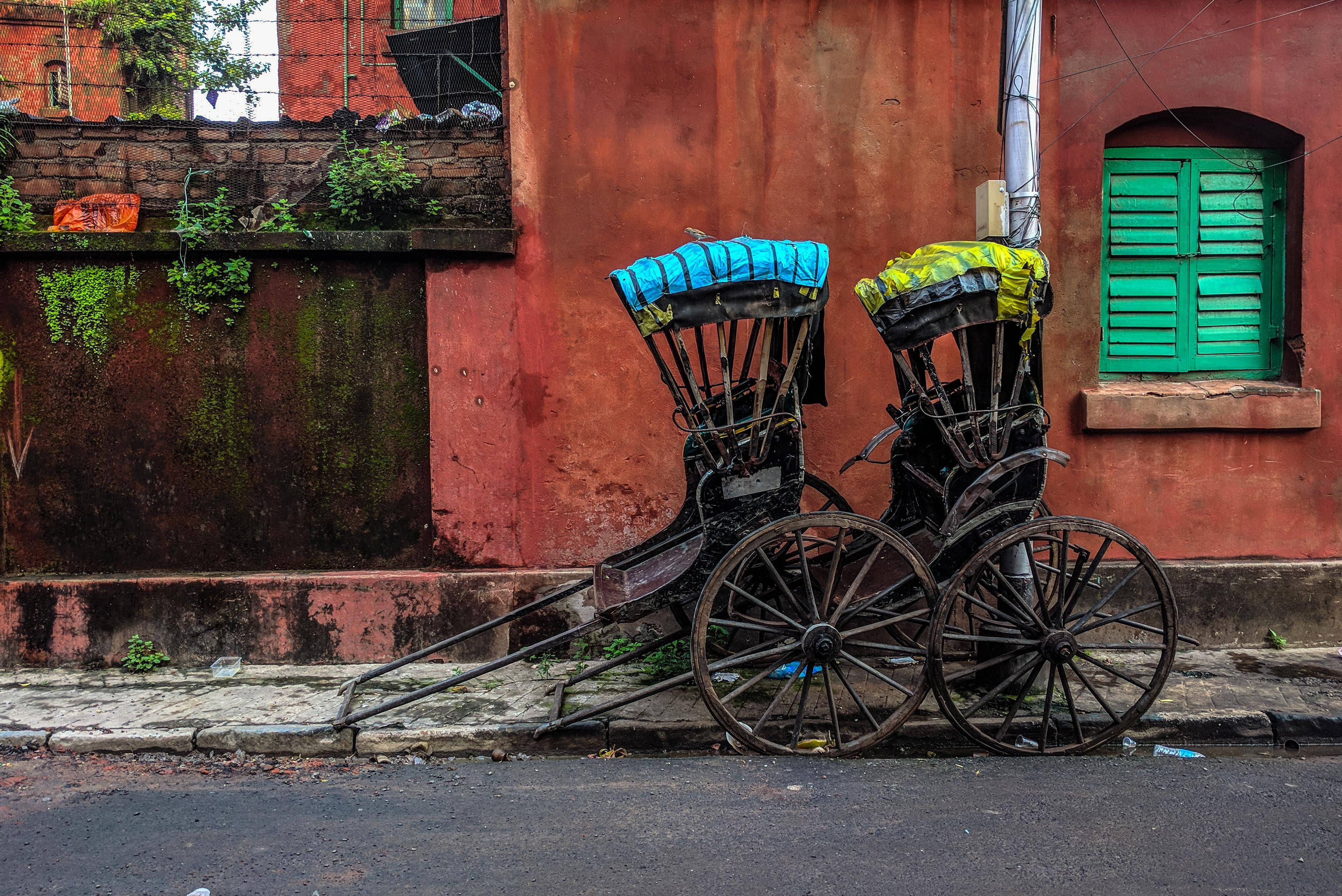 Kolkata 桌布 壁紙 — 兩輛風化的黃包車，帶有藍色和黃色頂篷，靠在一面鏽紅色的牆上，旁邊是一扇綠色百葉窗。