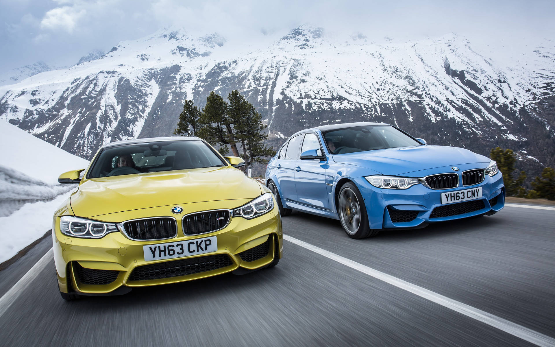 兩輛BMW跑車在白雪覆蓋的山峰背景下，於山路上飛馳。