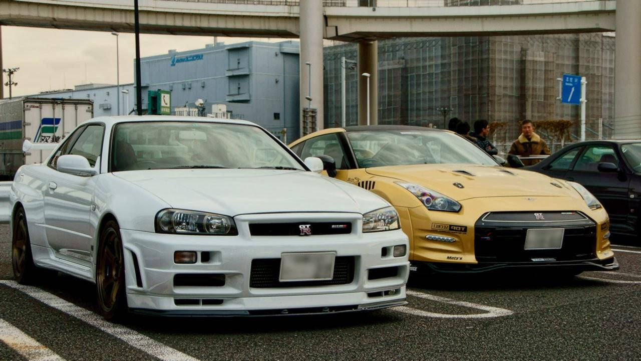 Road 桌布 壁紙 — 兩輛高性能日產Skyline GT-R，白色和金色，停在天橋下，背景是城市建築。