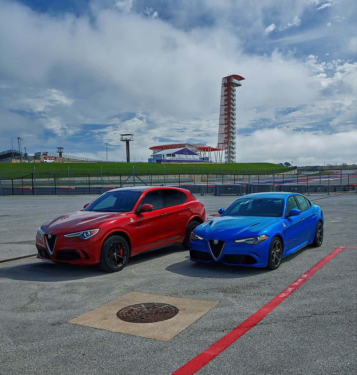 Alfa Romeo Stelvio 桌布 壁紙 — 兩輛鮮豔的紅色和藍色愛快羅密歐跑車停泊在賽道上，背景是陰天和一座塔。