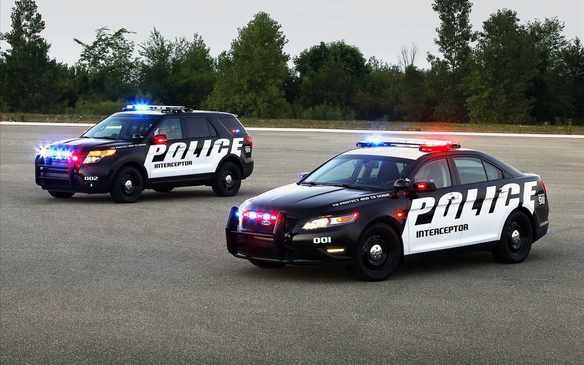 Police Car 桌布 壁紙 — 兩輛黑白警用攔截車閃著燈停泊在樹木環繞的瀝青停車場上。