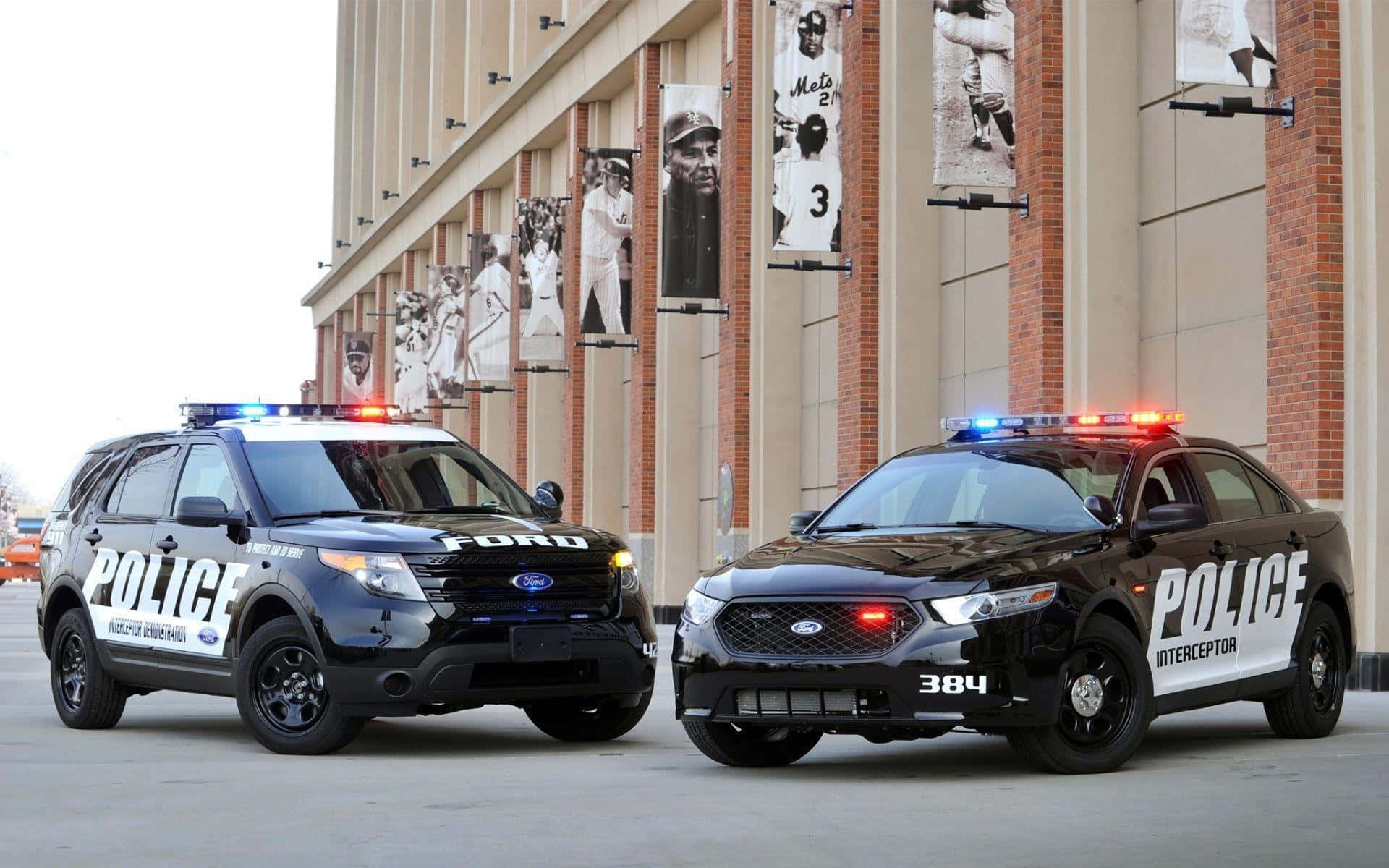 警察 桌布 壁紙 — 兩輛黑色福特警車閃著燈停放在一個掛有棒球選手橫幅的體育館外。