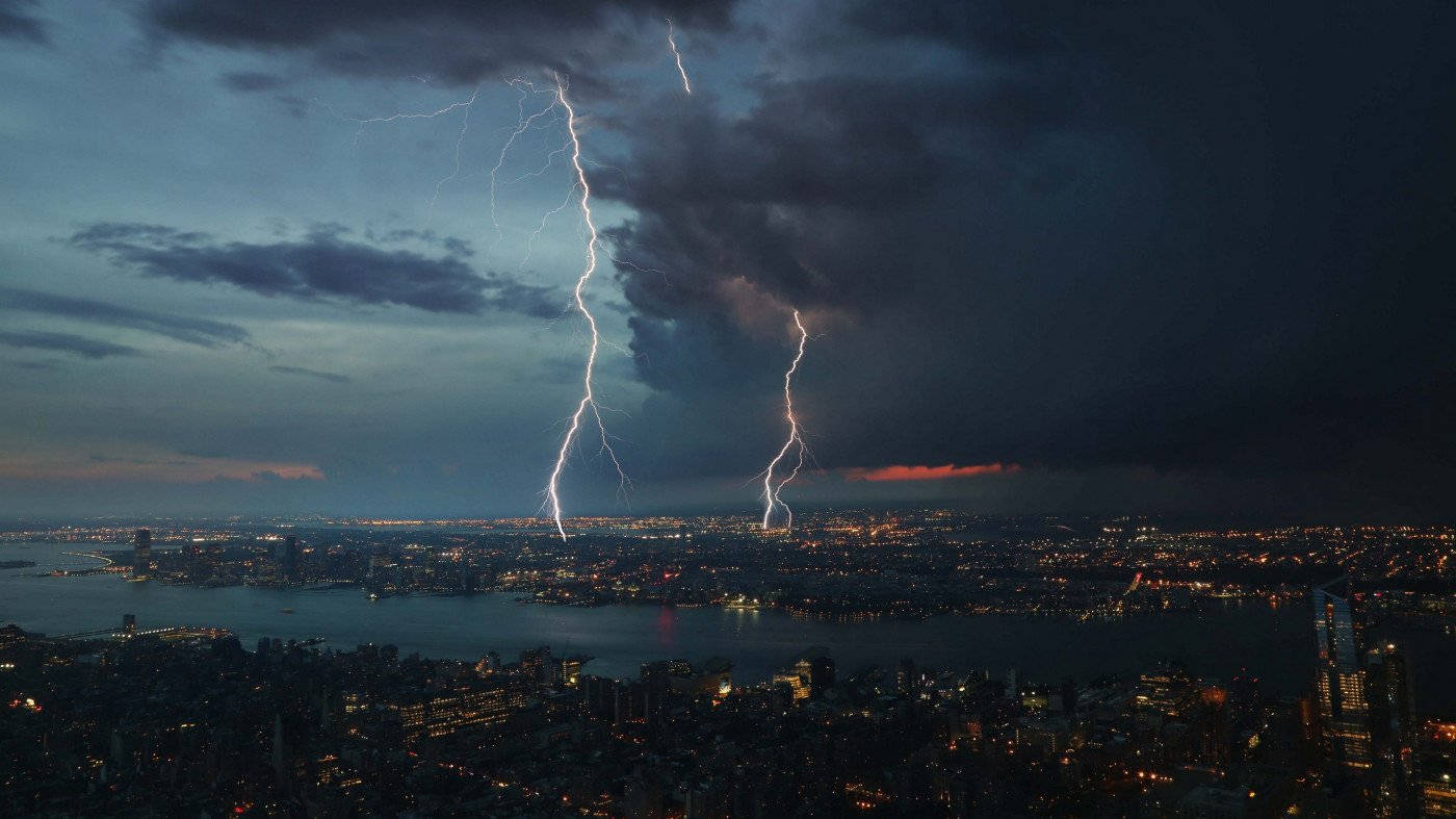 閃電 桌布 壁紙 — 兩道閃電劃破暴風雨中的暮色天空，照亮了下方無數窗戶閃爍的廣闊城市景觀。