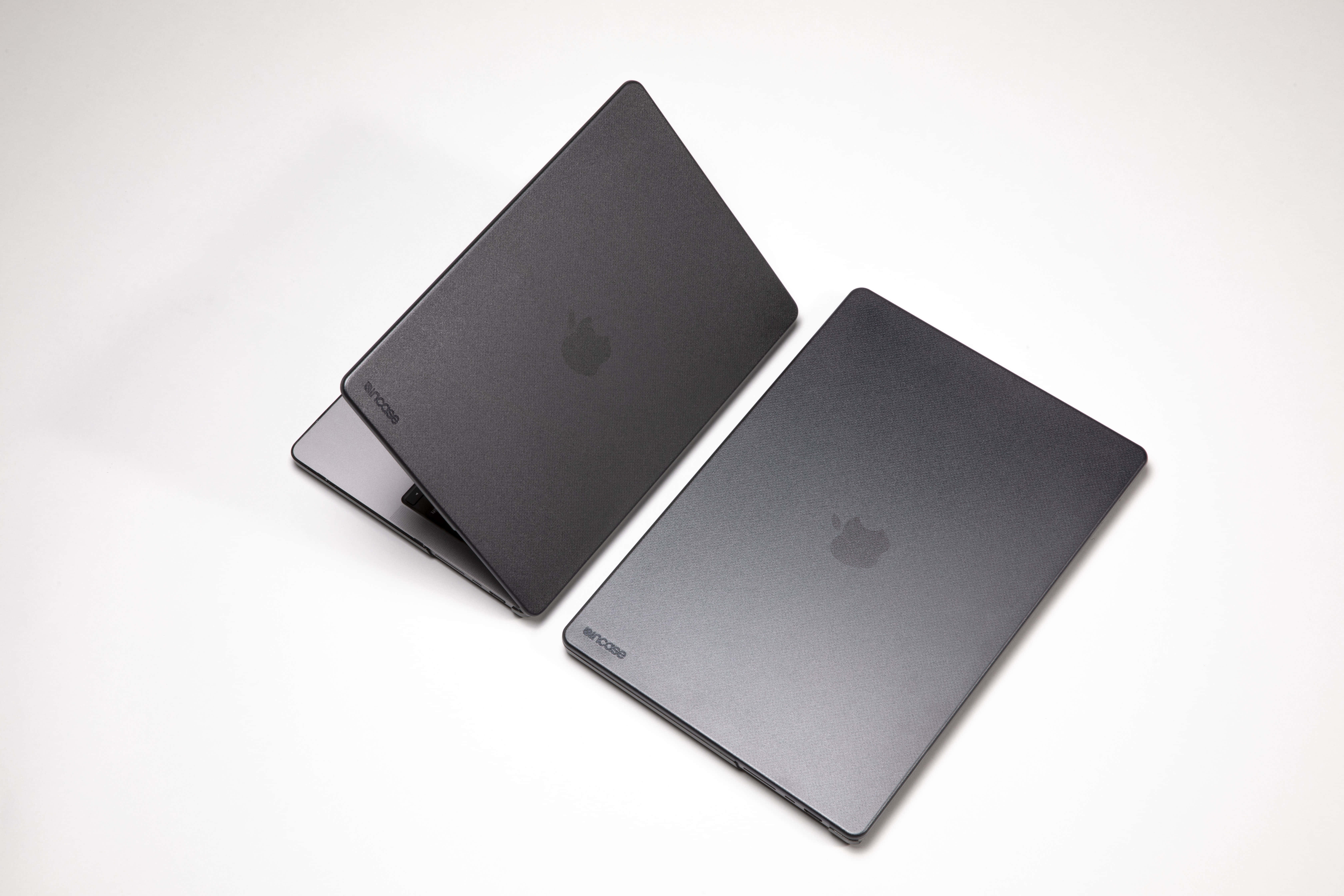 Black Macbook 桌布 壁紙 — 兩部帶有Apple標誌的霧面黑色MacBook筆記型電腦，在柔和的攝影棚燈光下傾斜放置在潔白的表面上。