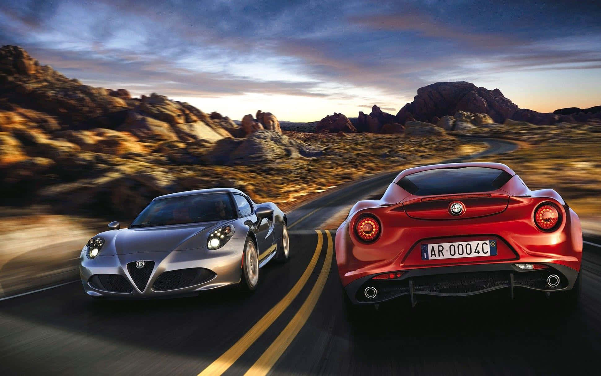 Alfa Romeo 4c 桌布 壁紙 — 兩部愛快羅密歐跑車在日落時分的沙漠公路上並排飛馳，背景是戲劇性的岩石地形。