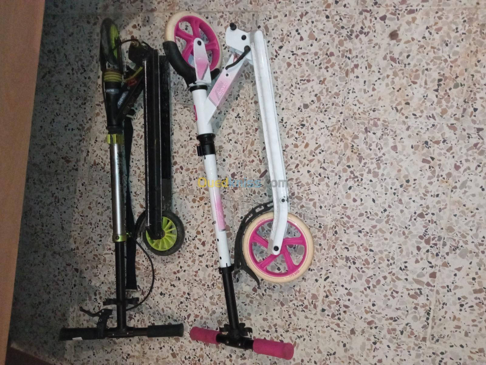 scooter 桌布 壁紙 — 兩部折疊滑板車，一部黑色配綠色車輪，另一部白色配粉紅色車輪，靠在一片斑點水磨石地板上。