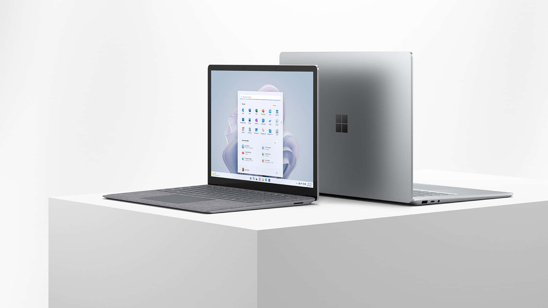 筆電 桌布 壁紙 — 兩部時尚的銀色 Microsoft Surface 筆記型電腦展示在白色底座上，一部打開顯示開始功能表，另一部關閉，極簡工作室拍攝。