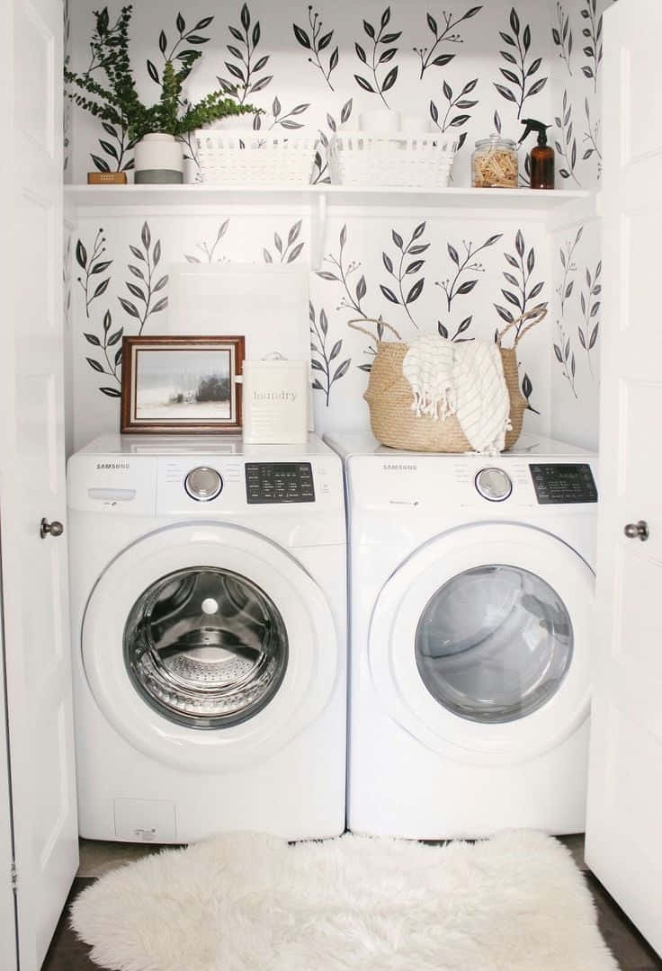 Laundry Room 桌布 壁紙 — 兩部白色前置式洗衣機並排放置在明亮的洗衣角落，配有葉子圖案壁紙和毛絨白色地毯。