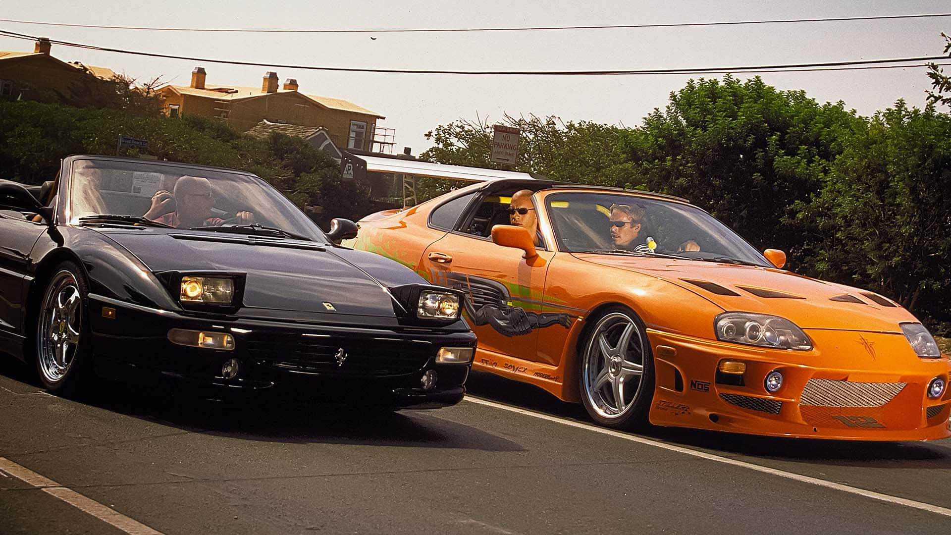 Fast And Furious 1 桌布 壁紙 — 兩部跑車，一部黑色法拉利和一部橙色豐田Supra，在陽光明媚的郊區街道上並排競速。