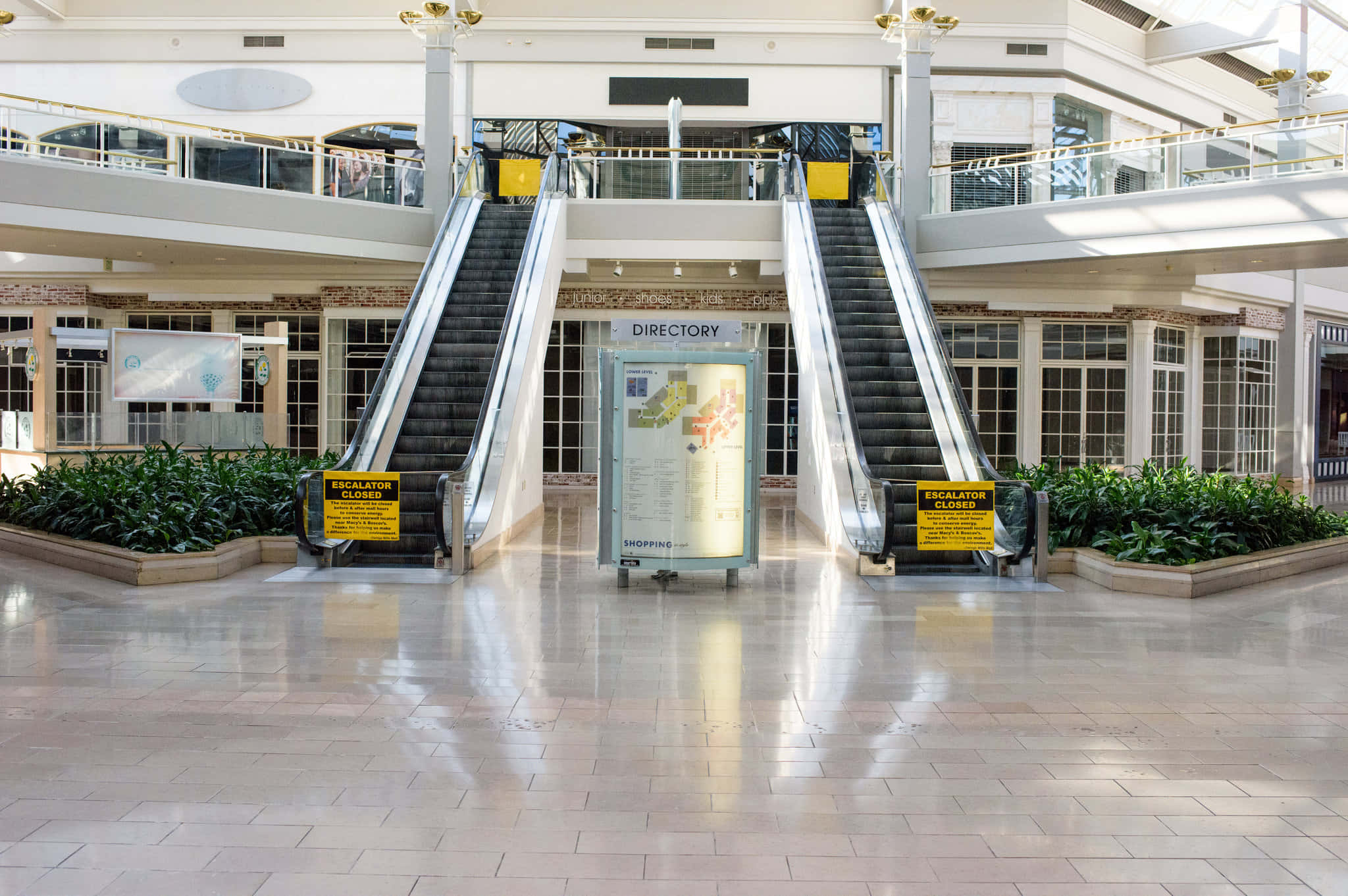 Mall 桌布 壁紙 — 兩部關閉的自動扶梯夾著一個目錄資訊亭，位於寬敞、陽光充足的購物中心中庭，舖有瓷磚地板並擺放盆栽植物。