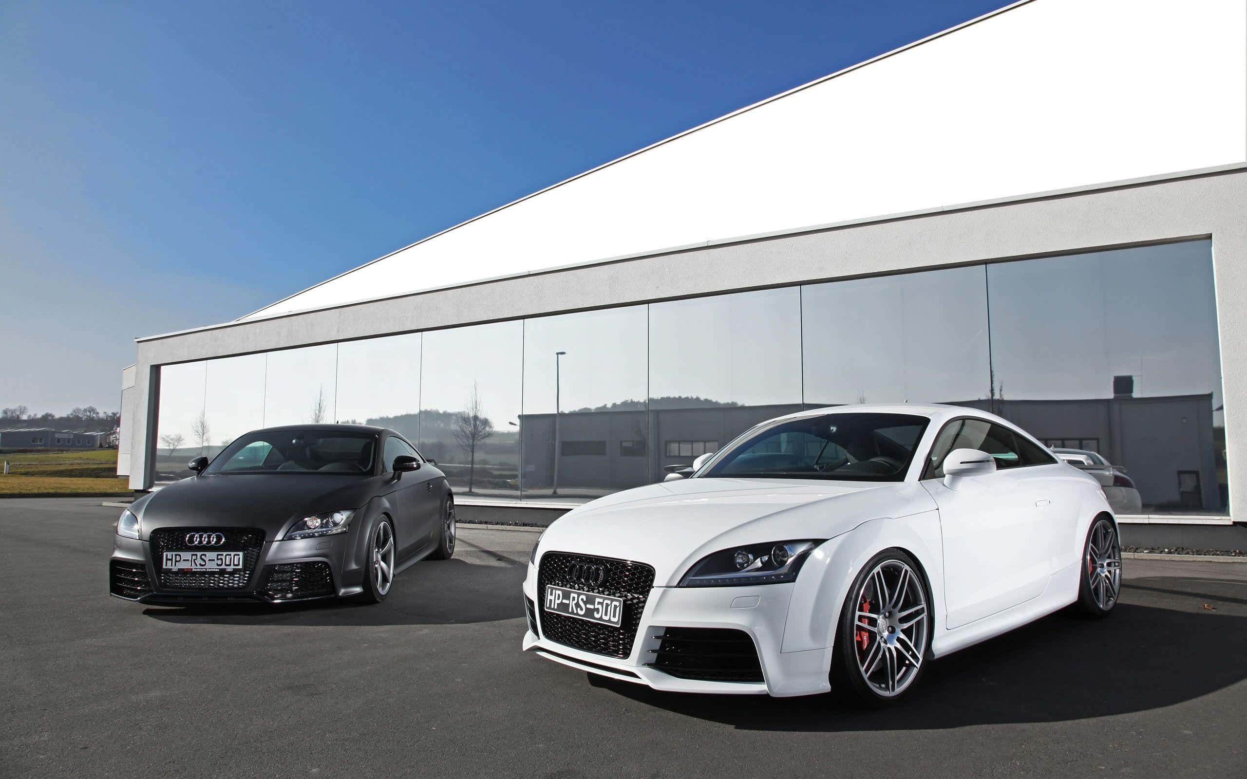 Audi Tt Rs 桌布 壁紙 — 兩部高性能奧迪 TT RS 500 雙門跑車，一部消光黑色，一部亮光白色，停放在現代玻璃建築前，天空清澈湛藍。