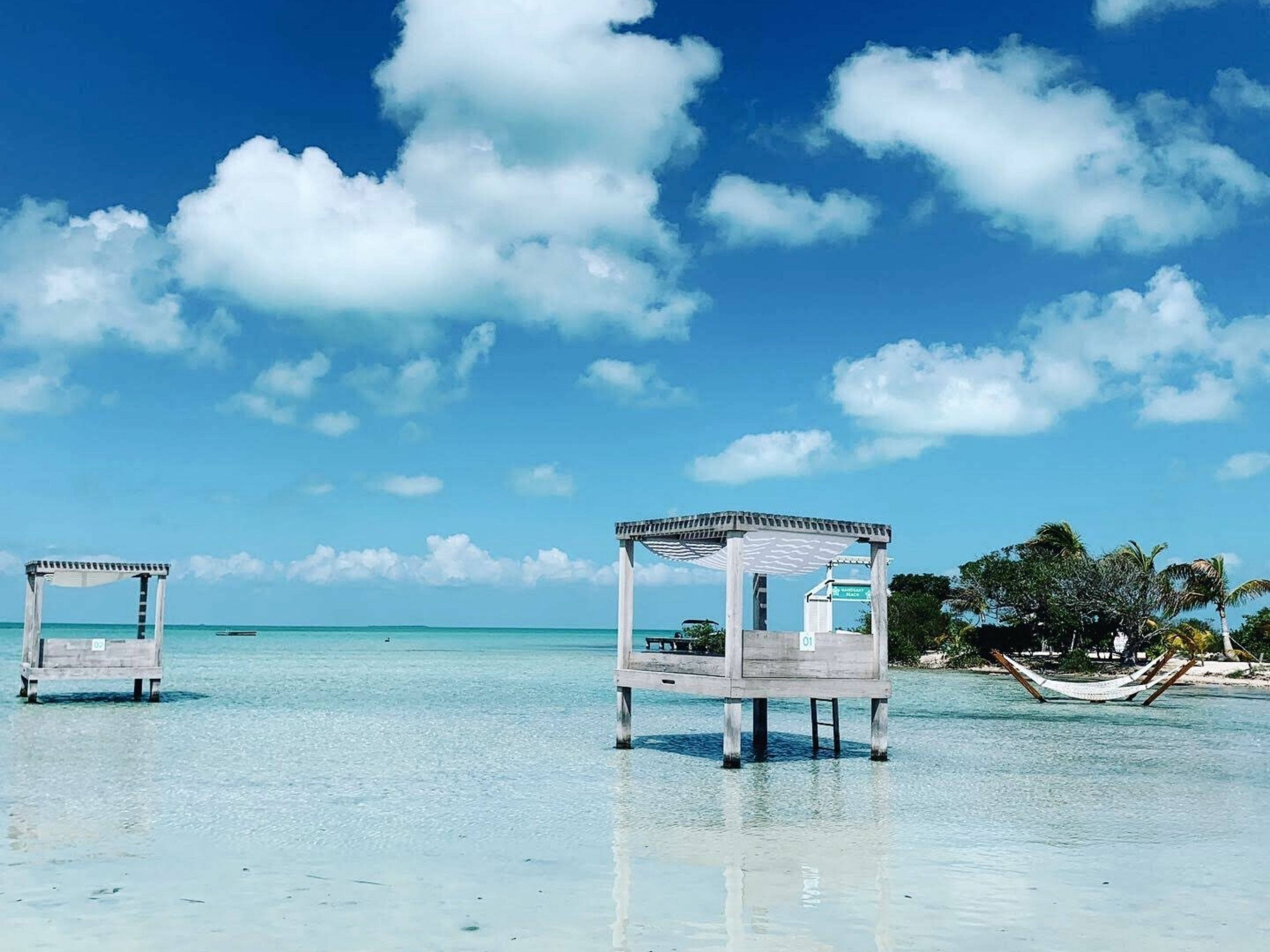 Belize 桌布 壁紙 — 兩間木製小屋漂浮在清澈碧綠的海水上，上方是明亮的藍天和蓬鬆的白雲。