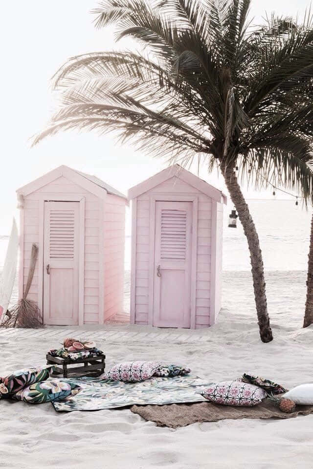 Beach Huts 桌布 壁紙 — 兩間粉彩粉紅色沙灘小屋矗立在沙灘上，下方有搖曳的棕櫚樹，附近有圖案靠墊和衝浪板。