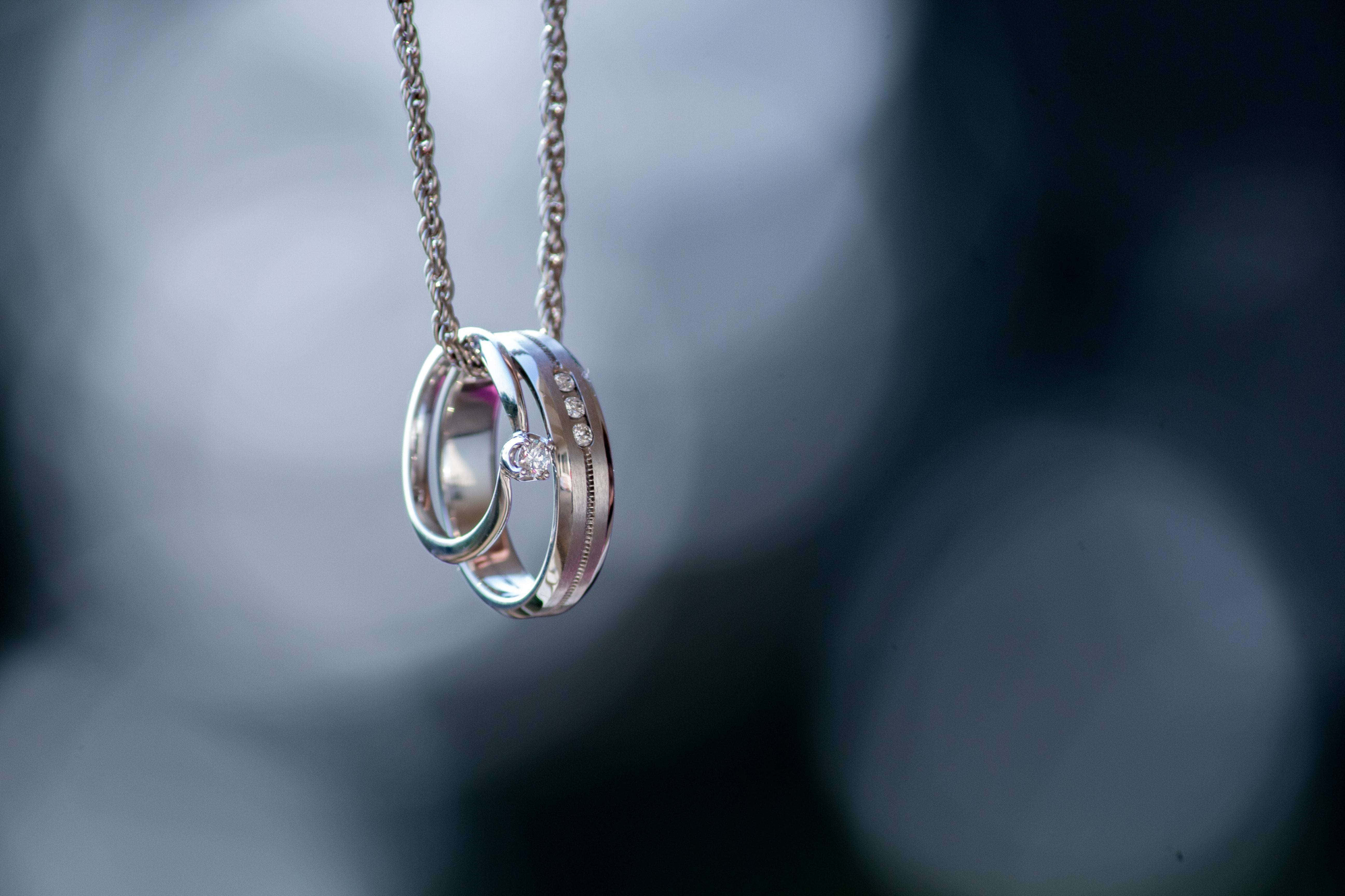 珠寶 桌布 壁紙 — 兩隻交織的銀色結婚戒指鑲有鑽石，從一條鏈條上精緻地懸掛在柔和模糊的灰色背景前。