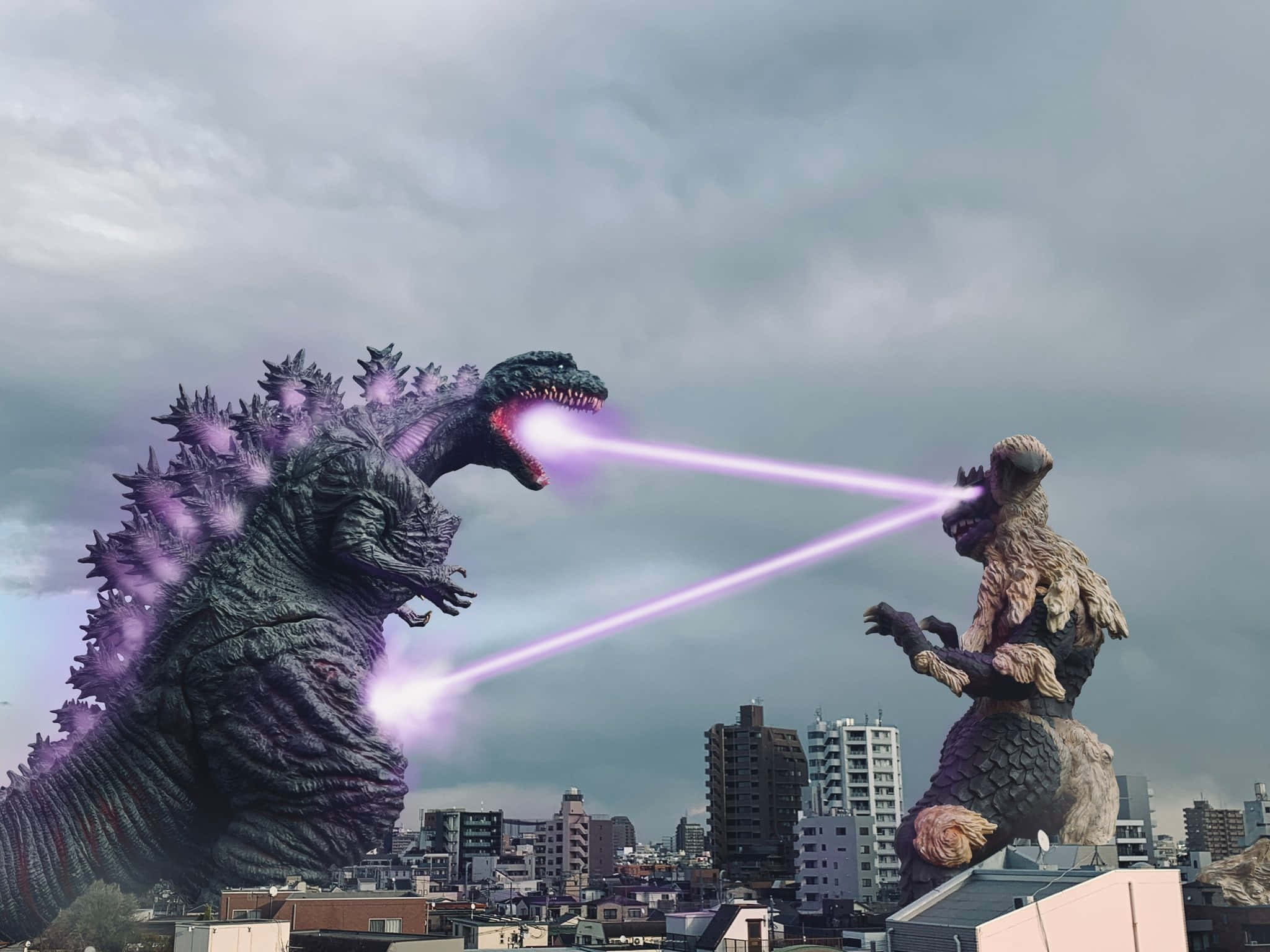Godzilla Vs King Caesar 桌布 壁紙 — 兩隻巨型怪獸哥吉拉和王者基多拉在城市天際線上戰鬥，向多雲的天空發射紫色原子吐息。