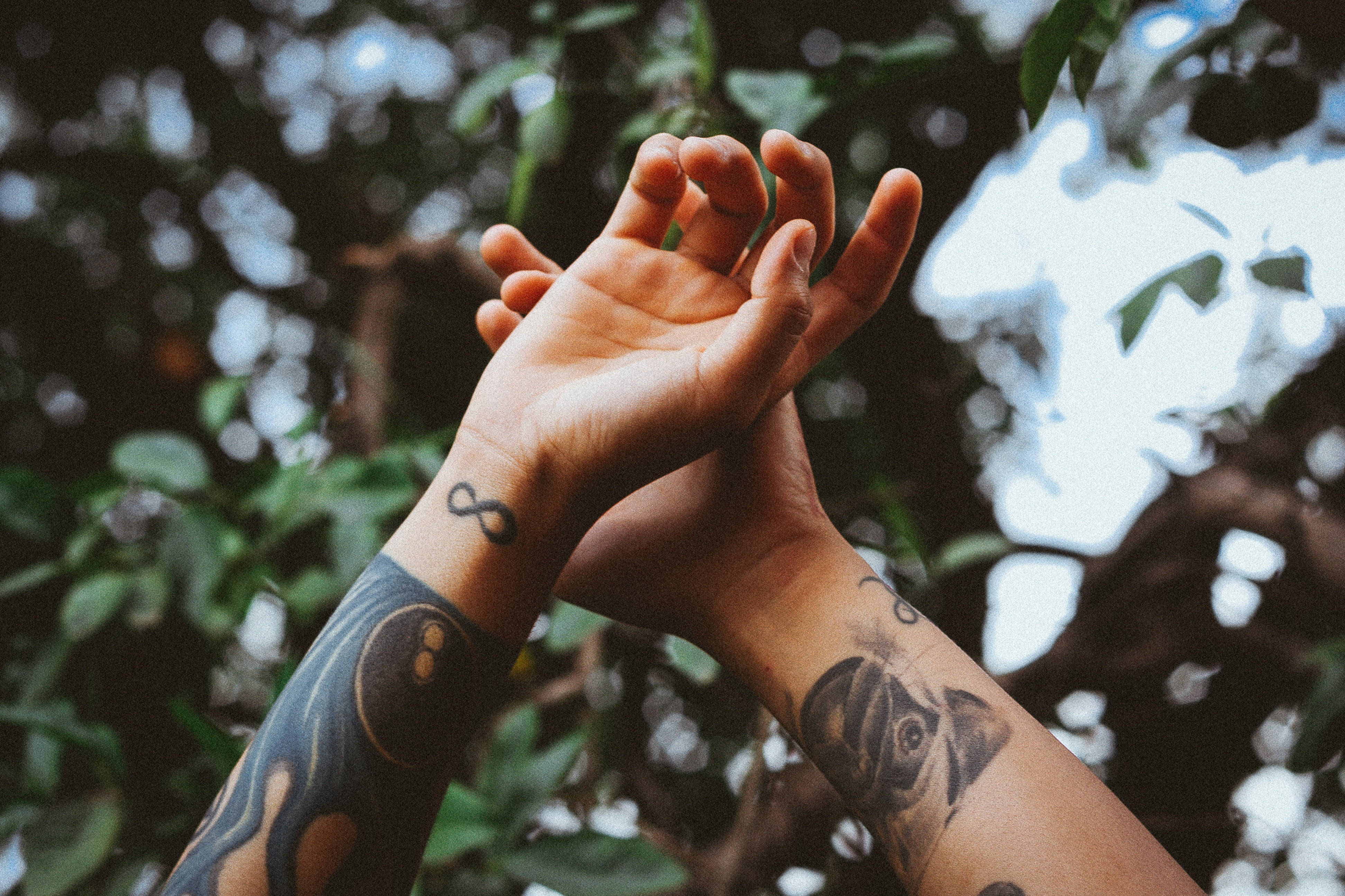 Tattoo 桌布 壁紙 — 兩隻帶有刺青的手輕輕交握，背景是柔和模糊的綠葉和斑駁的陽光。