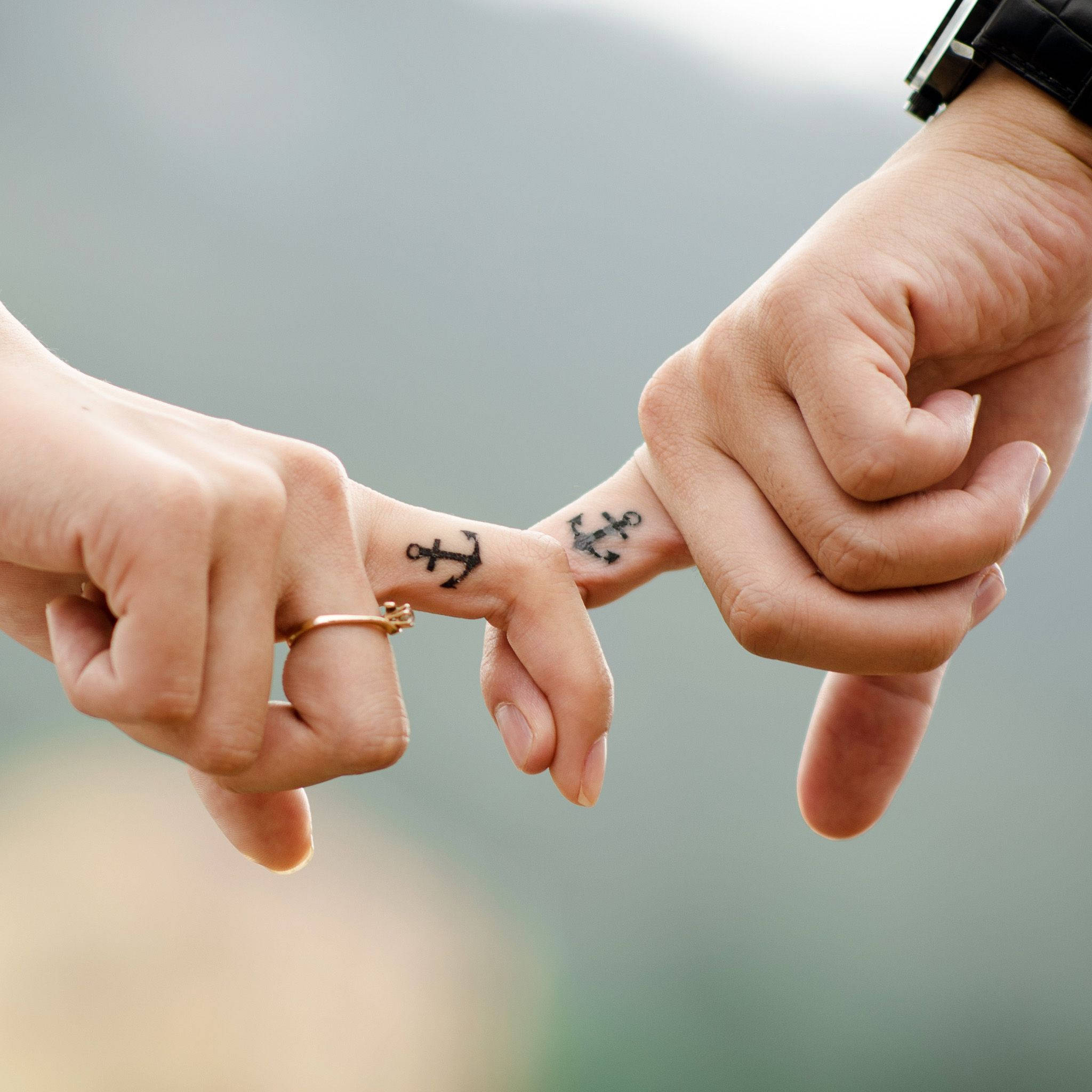 Aesthetic Love 桌布 壁紙 — 兩隻帶有匹配船錨紋身的手，在柔和模糊的山景背景下輕輕觸碰指尖。