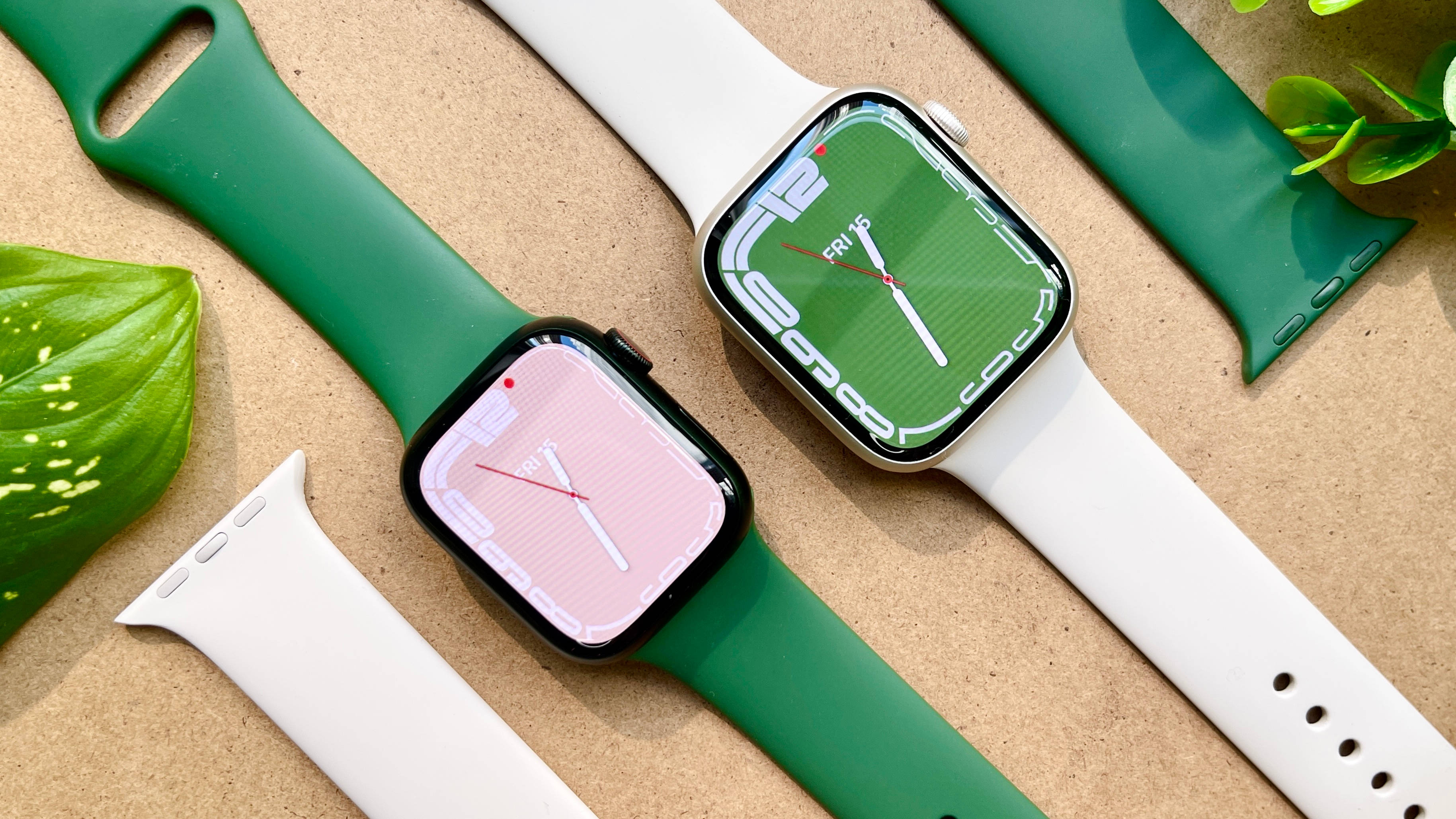 Apple Watch 動態 桌布 壁紙 — 兩隻帶有綠色和白色錶帶的智慧手錶放在有紋理的紙板上，旁邊是茂盛的綠葉。
