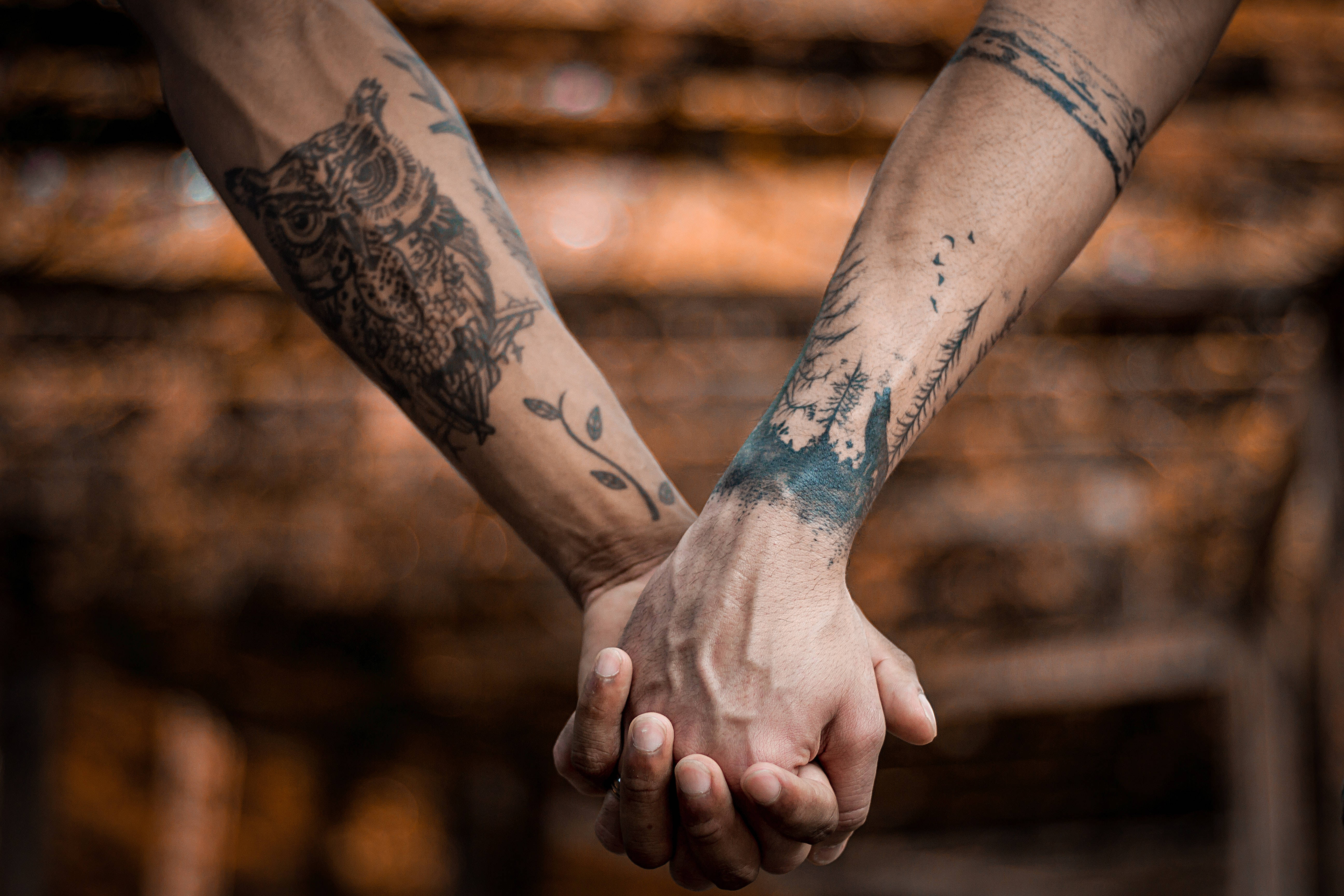 Tattoo 桌布 壁紙 — 兩隻帶有貓頭鷹和森林圖案紋身的手臂，在溫暖模糊的木質背景前緊緊握著手。