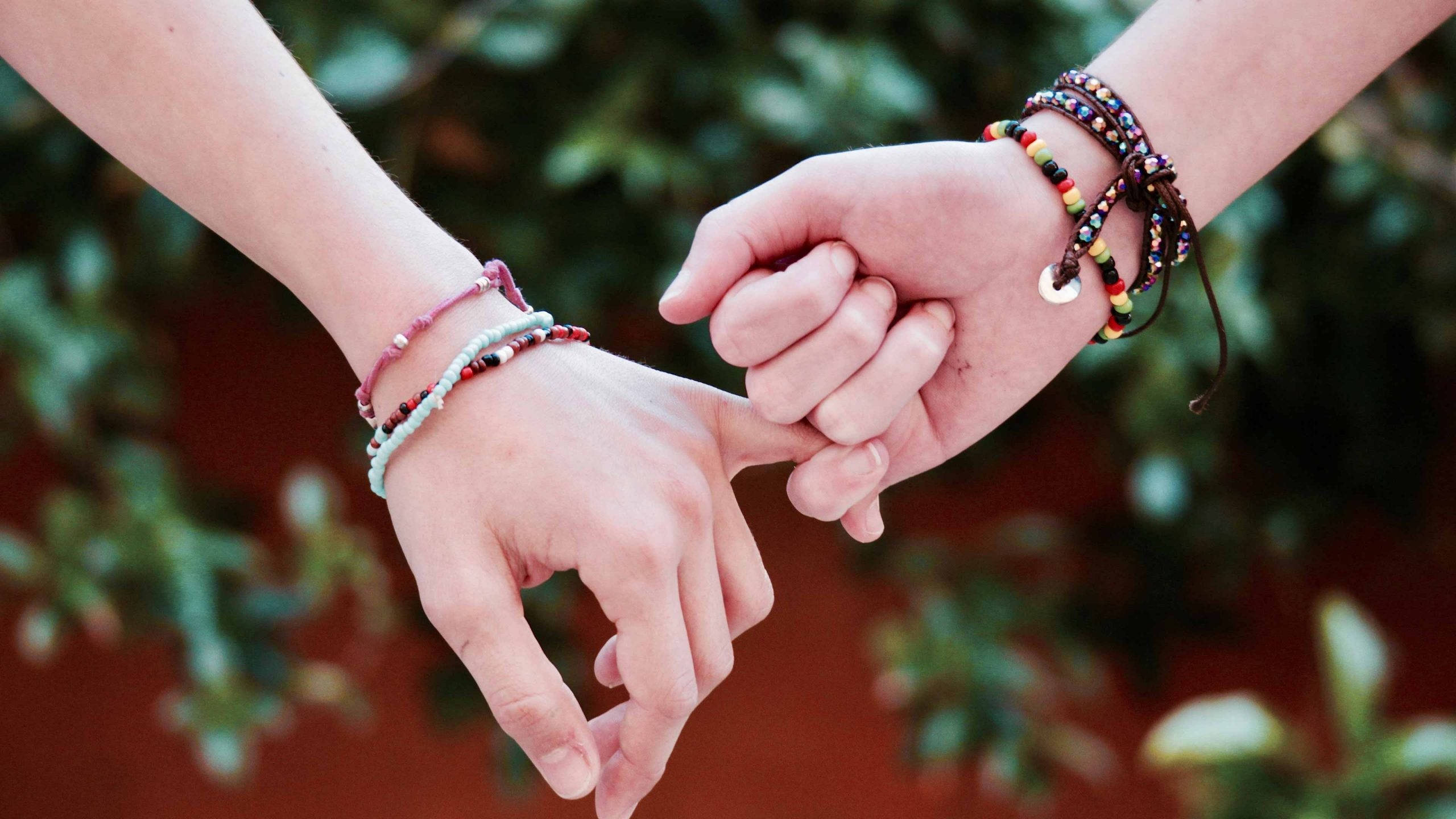 Friendship 桌布 壁紙 — 兩隻戴著彩色珠串手鐲的手在模糊的綠色和紅色背景上形成一個心形。