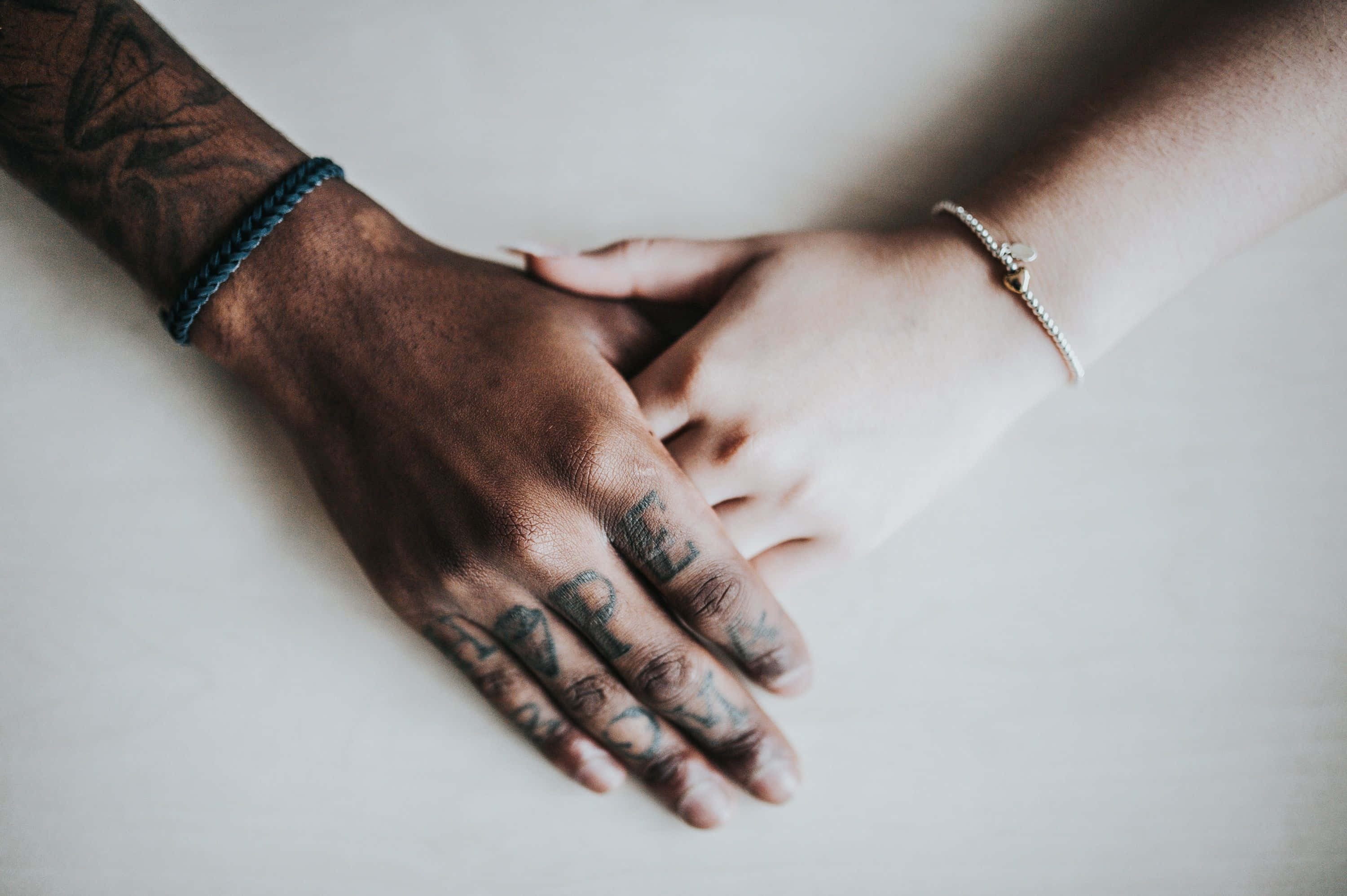 Interracial 桌布 壁紙 — 兩隻手，一隻有刺青和藍色繩子，另一隻有銀色手鐲，輕輕地握在一起，放在一個平坦的表面上。