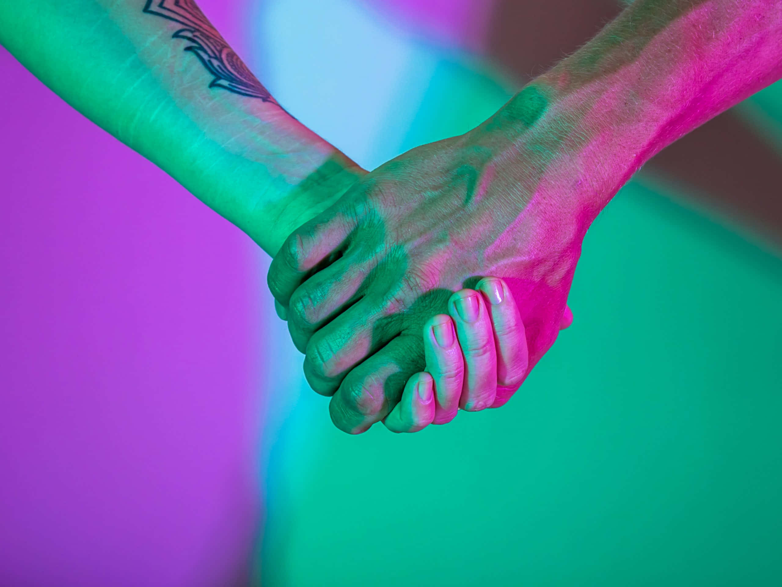 Holding Hands 桌布 壁紙 — 兩隻手在充滿活力的霓虹粉紅和綠色燈光下緊緊相握，一隻手臂上可見刺青。