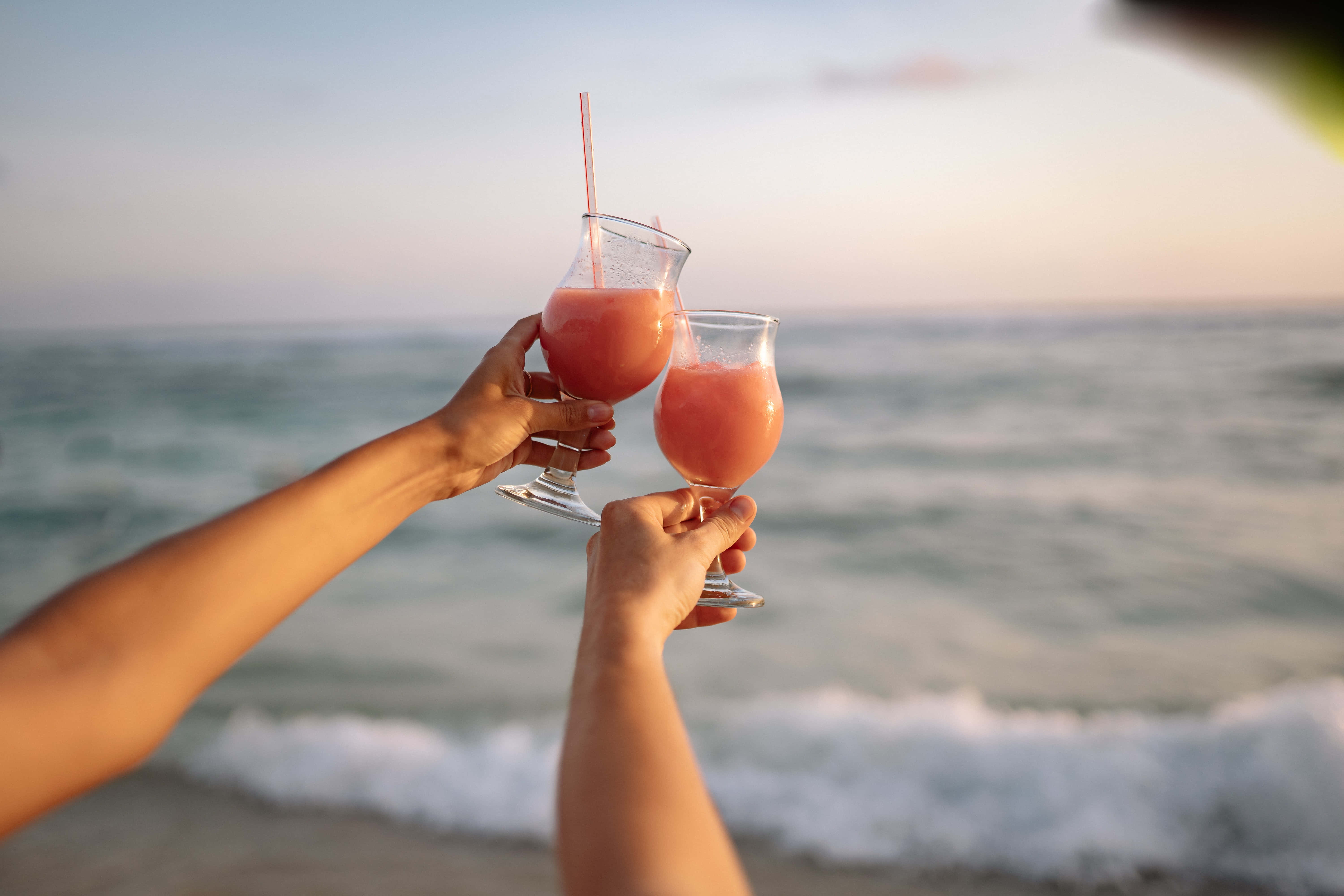Summer Drinks 桌布 壁紙 — 兩隻手在日落時分的沙灘上，隨著海浪翻滾，碰杯粉紅色的飲料。