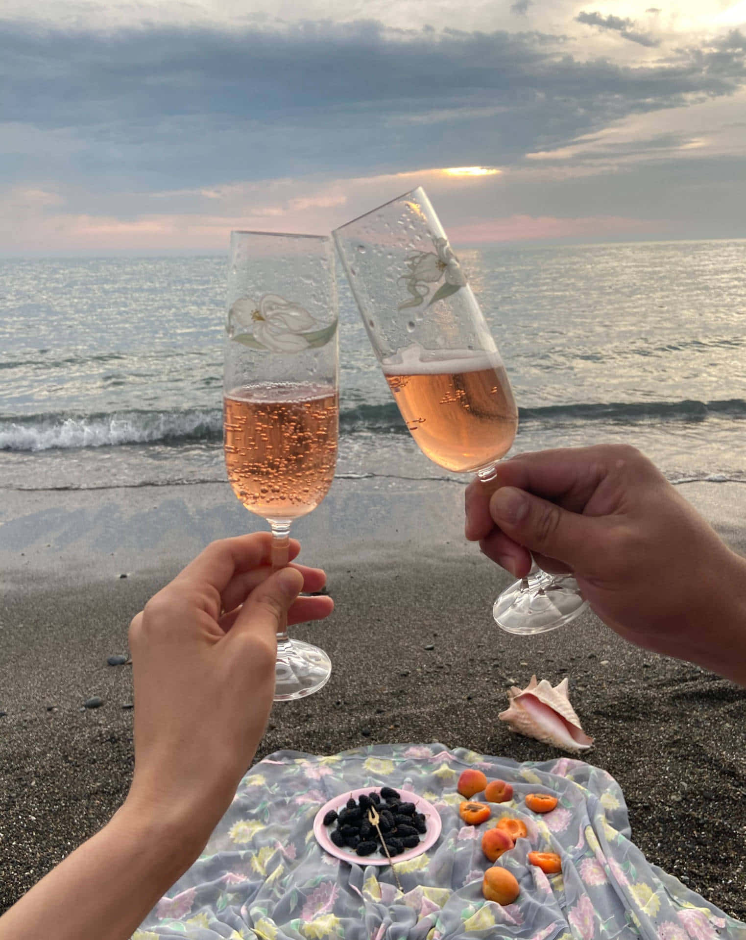 Beach Picnic 桌布 壁紙 — 兩隻手在日落時分的沙灘上碰香檳杯，附近有圖案毯子和水果。