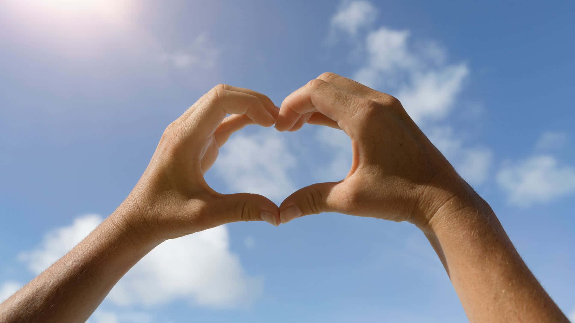 Beautiful Love 桌布 壁紙 — 兩隻手在明亮的藍天中形成心形，天空中有散落的白雲和太陽耀斑。