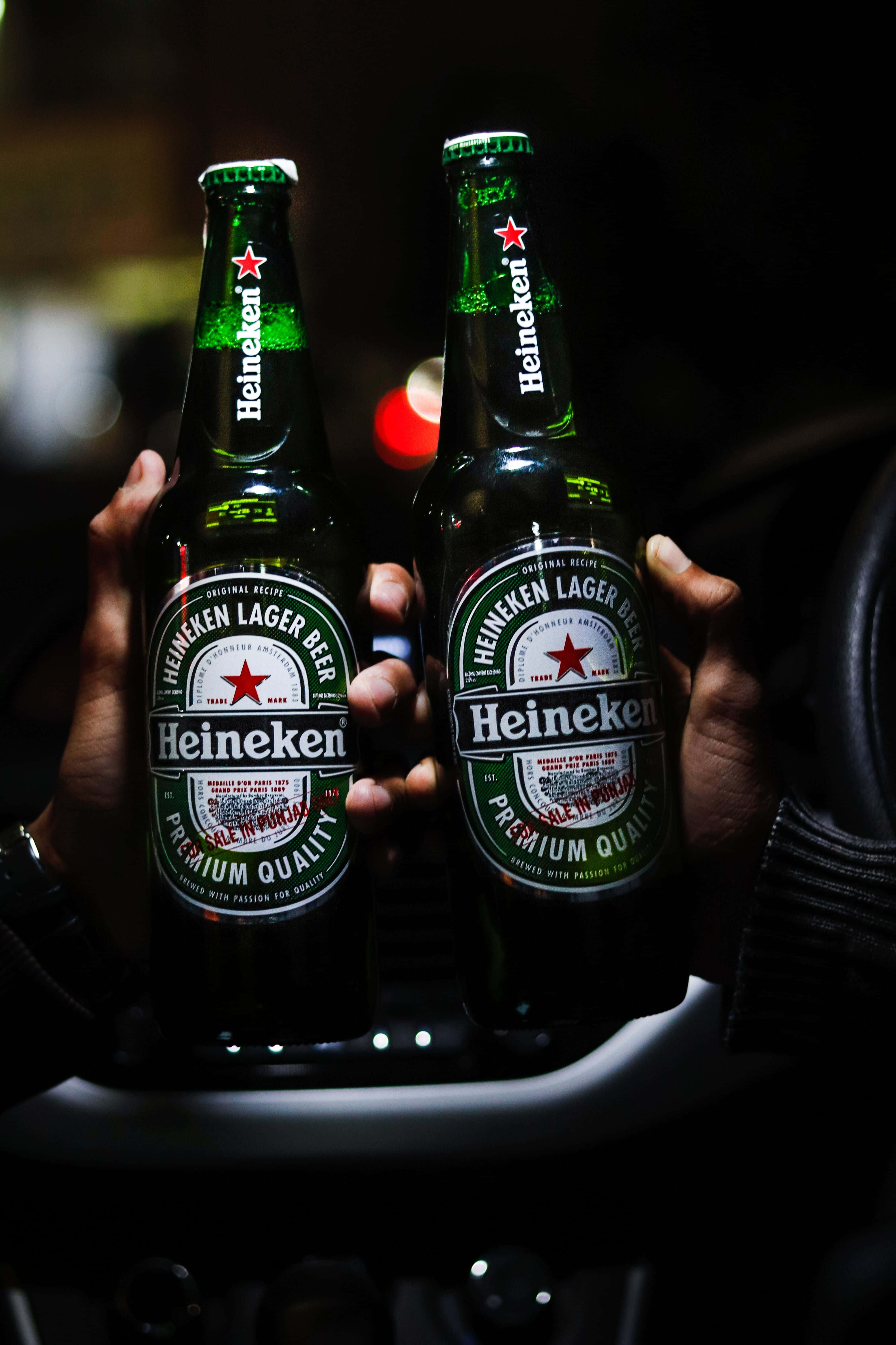 Heineken 桌布 壁紙 — 兩隻手在昏暗的車廂內握著綠色海尼根啤酒瓶，背景有模糊的燈光。