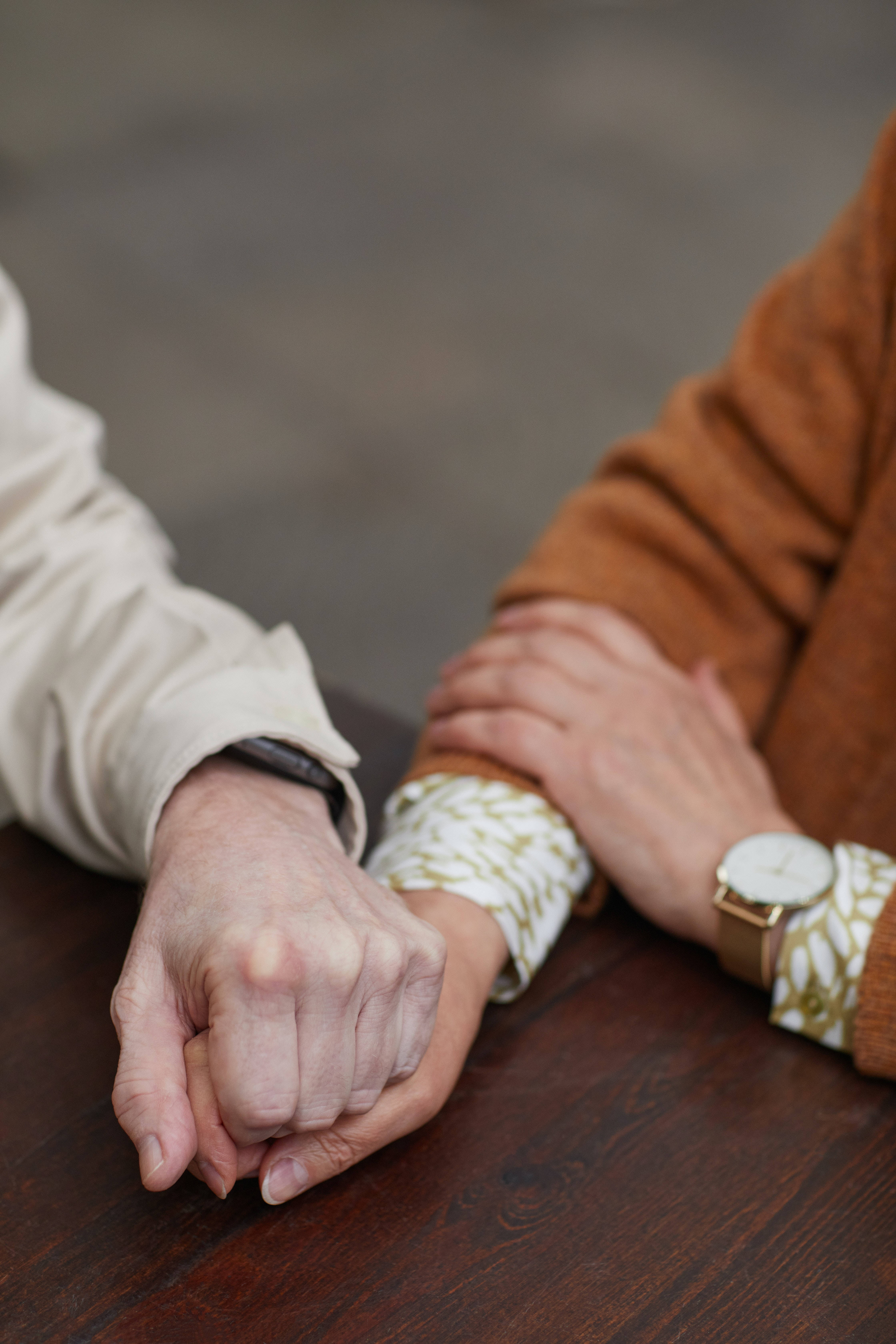 Couple Hands 桌布 壁紙 — 兩隻手在木桌上緊握，一隻戴著手錶和圖案袖，另一隻戴著黑色腕帶。