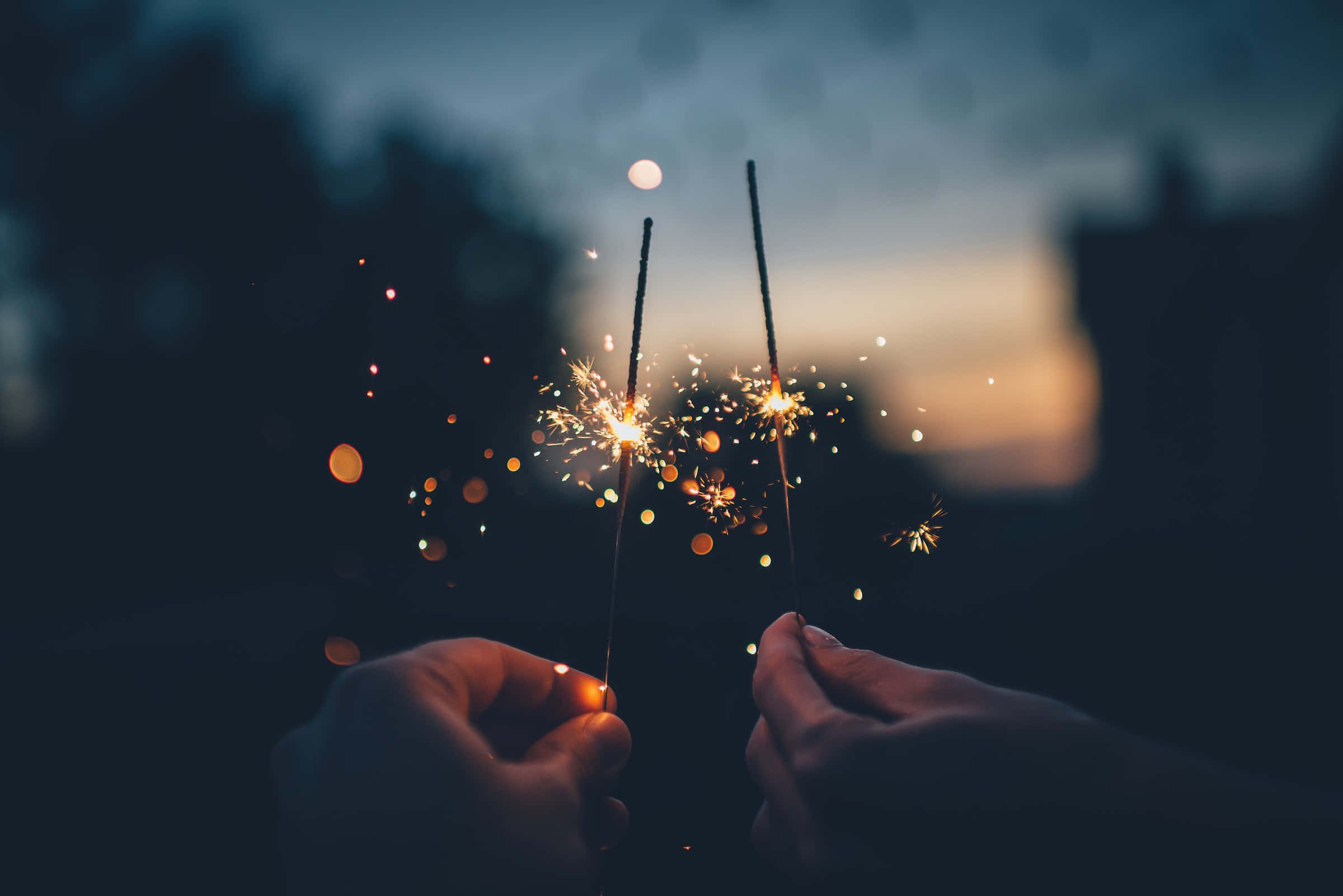 sparklers 桌布 壁紙 — 兩隻手在柔和散景燈光的暮色天空下，舉起閃閃發光的煙火。