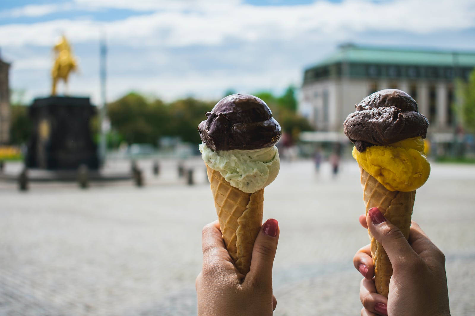 Ice Cream 桌布 壁紙 — 兩隻手在模糊的城市廣場背景（有金色雕像）前，拿著巧克力和香草冰淇淋甜筒。
