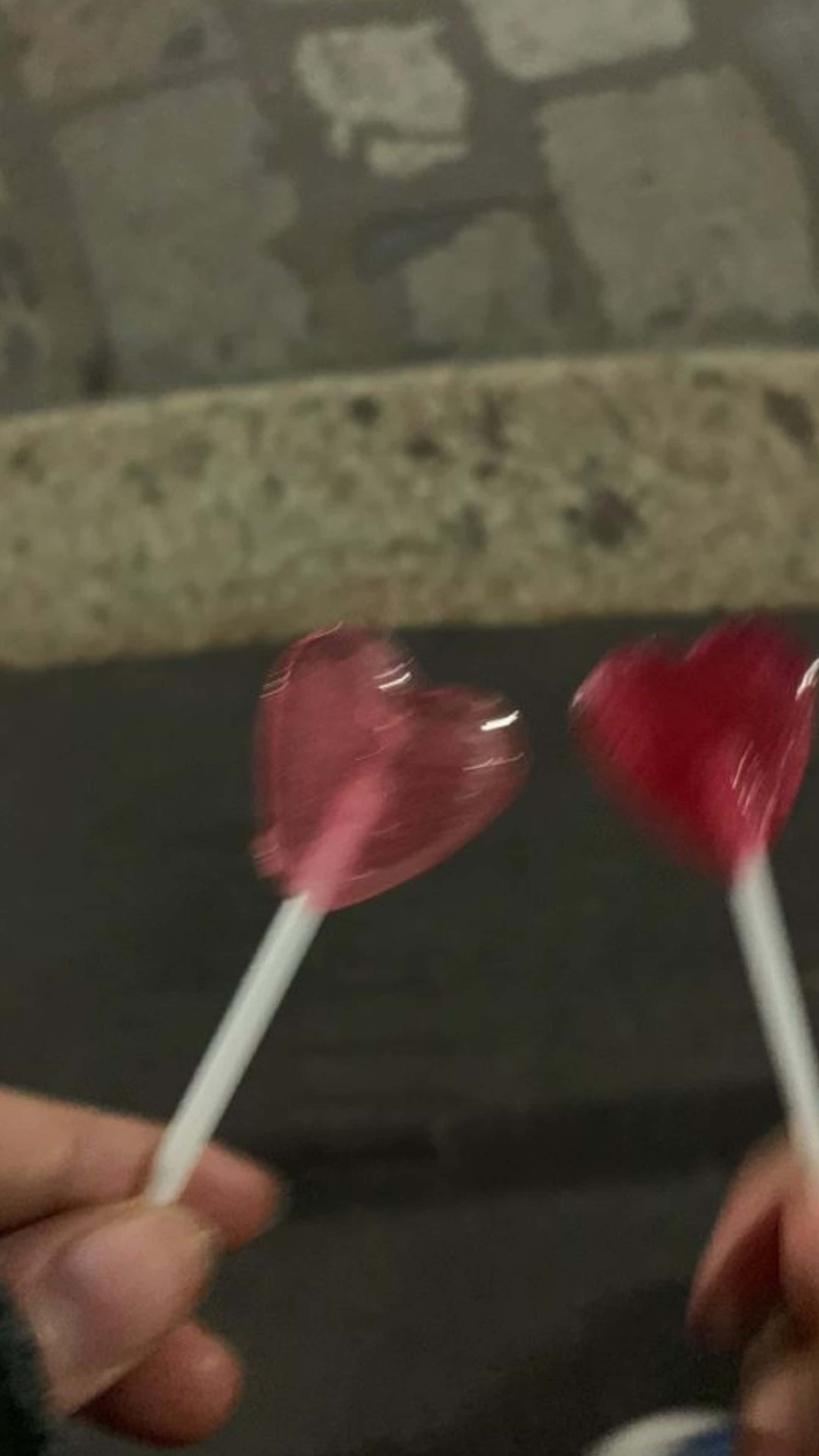 Red Hearts 桌布 壁紙 — 兩隻手在模糊的石鋪背景前拿著心形粉紅色棒棒糖。
