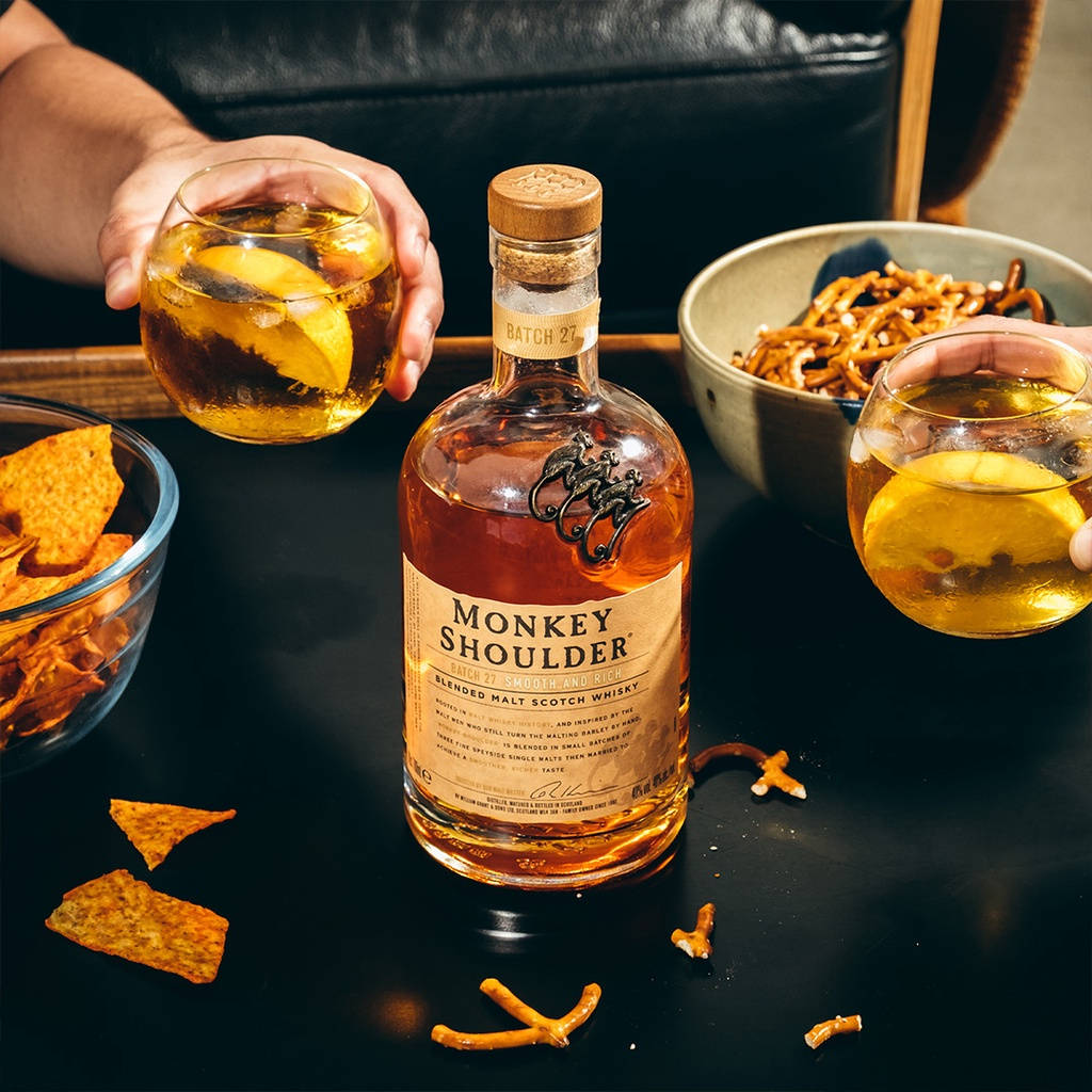 alcohol 桌布 壁紙 — 兩隻手在深色桌子上，旁邊放著一瓶Monkey Shoulder和裝有椒鹽捲餅和薯片的碗，舉起帶有檸檬片的威士忌酒杯碰杯。