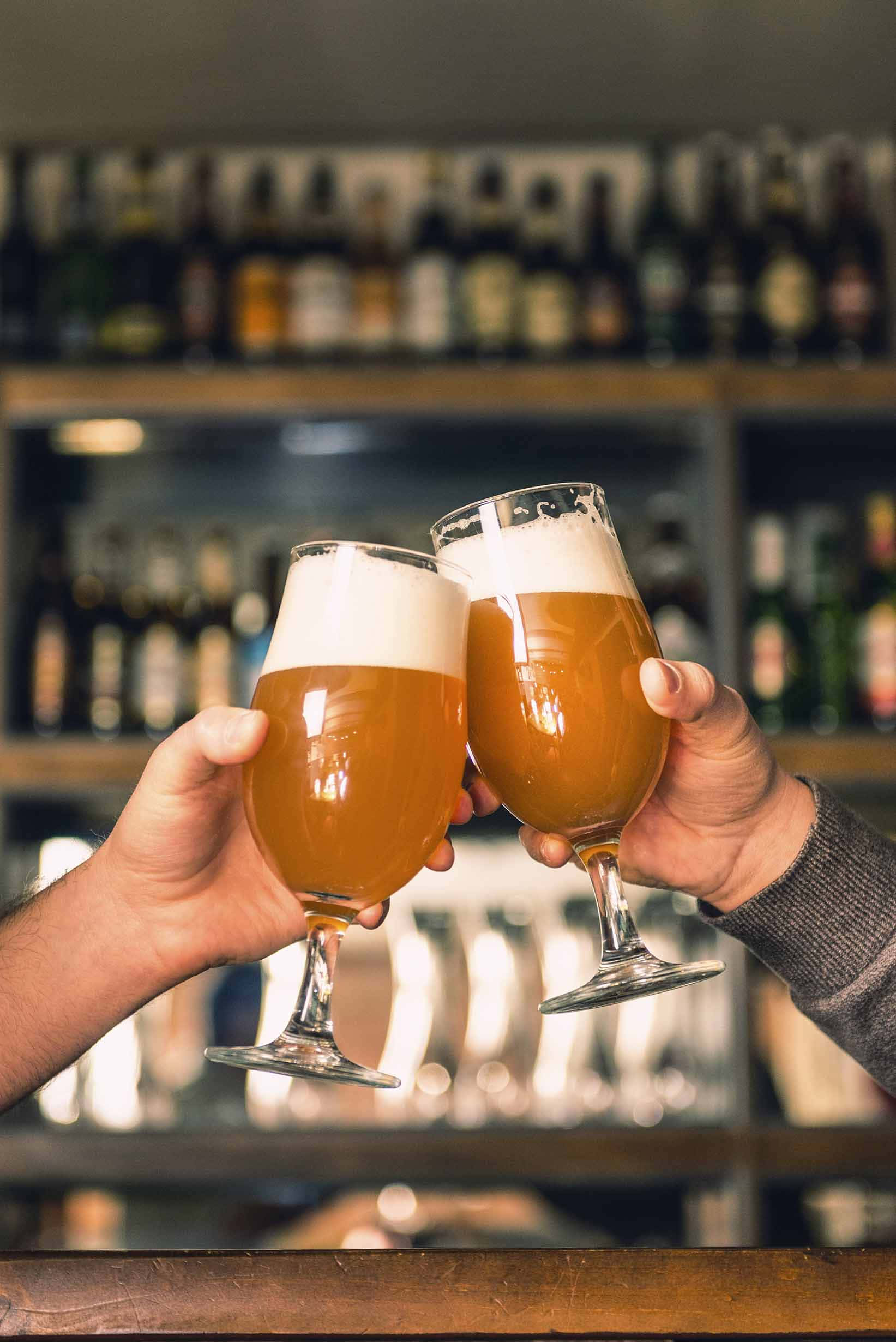 Beer 桌布 壁紙 — 兩隻手在溫暖燈光的酒吧中碰杯，琥珀色啤酒杯帶有泡沫，背後是模糊的酒架。