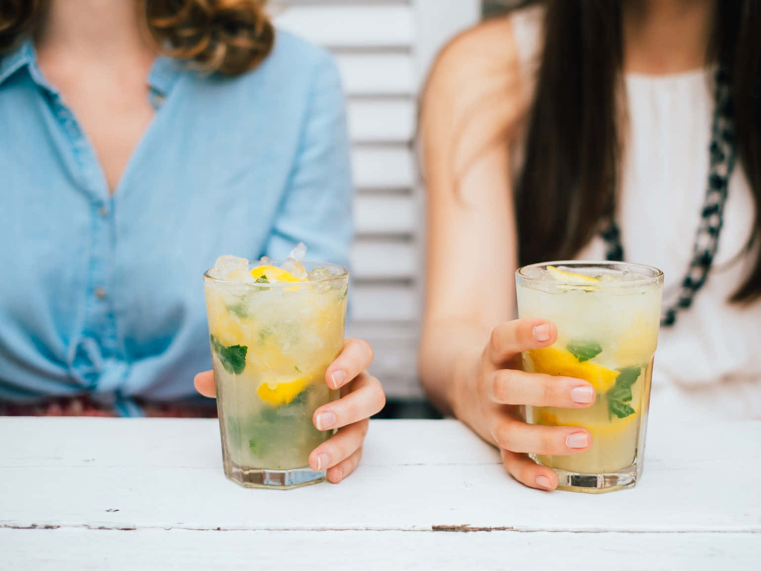 Beautiful Drink 桌布 壁紙 — 兩隻手在白色木桌上拿著清新的檸檬薄荷飲品，背景有模糊的人影。