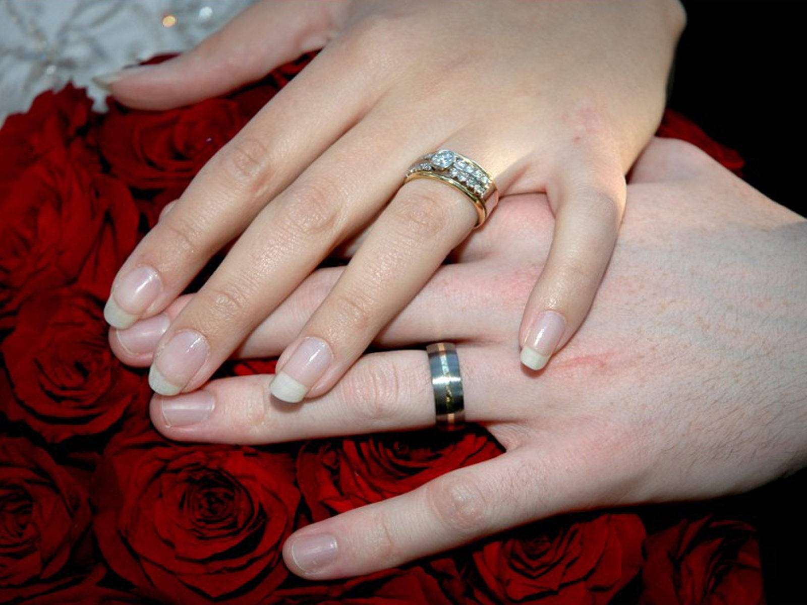 Wedding Rings 桌布 壁紙 — 兩隻手在紅玫瑰上交握，一隻戴著鑽石訂婚戒指，另一隻戴著簡單的結婚戒指。