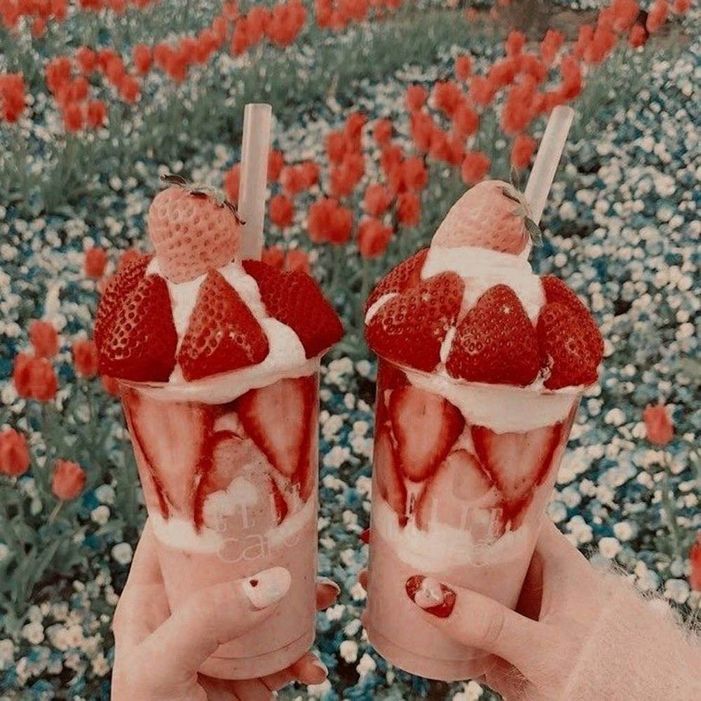 Strawberry Aesthetic 桌布 壁紙 — 兩隻手在紅白花田背景下，拿著頂部有新鮮草莓的草莓奶昔。