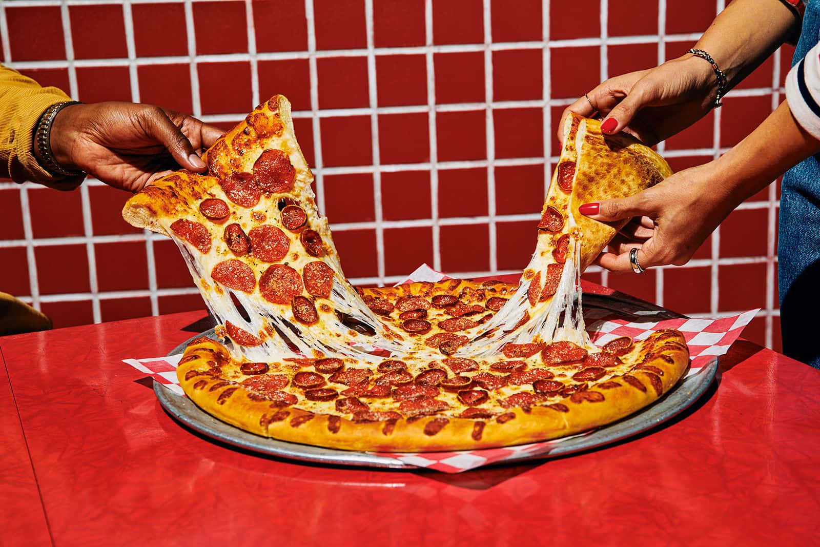 Pizza 桌布 壁紙 — 兩隻手在紅色瓷磚檯面上拉開一塊起司辣肉腸披薩，起司拉絲效果戲劇性。