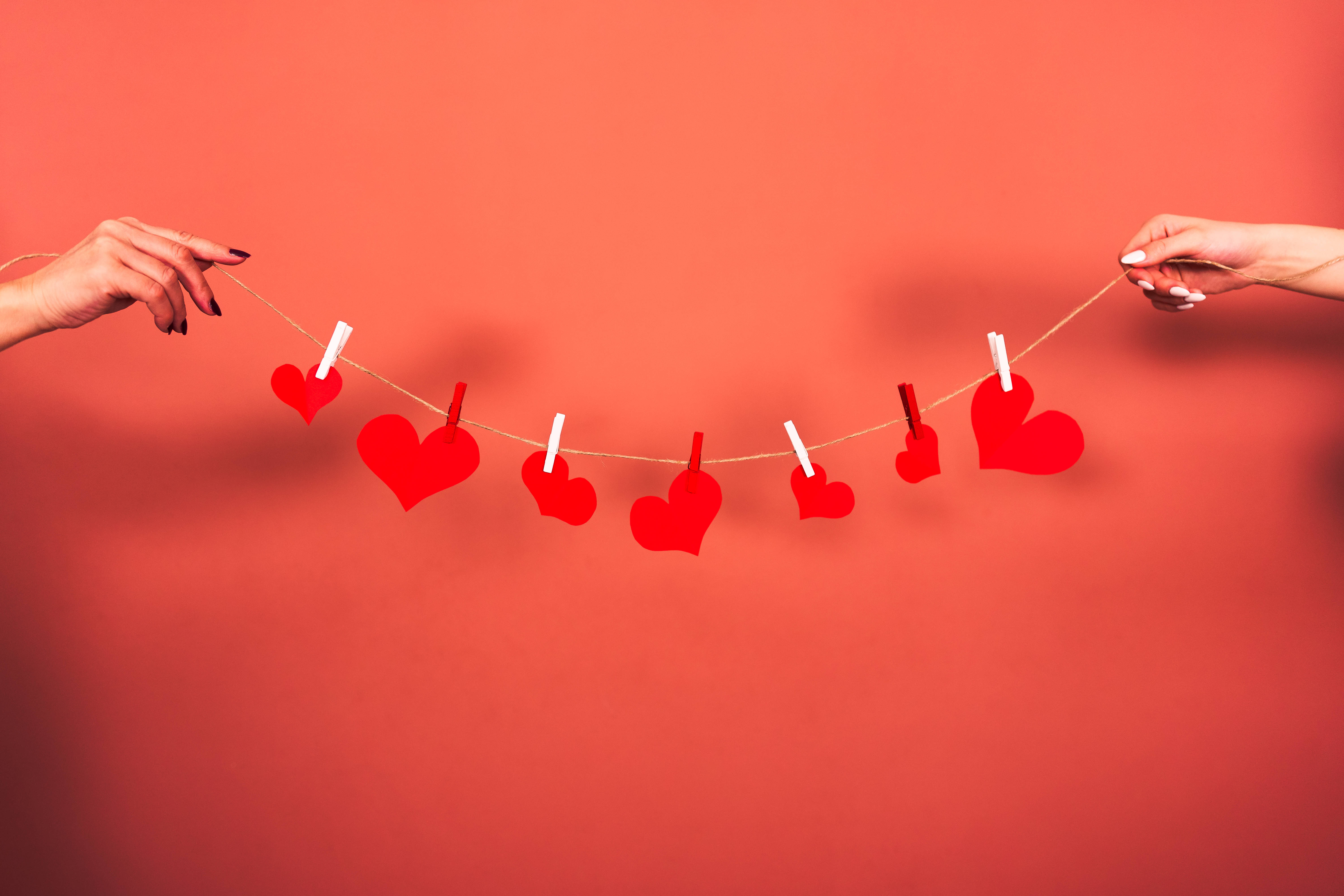 Aesthetic Heart 桌布 壁紙 — 兩隻手在純珊瑚色背景前拉開一串紅色紙愛心。