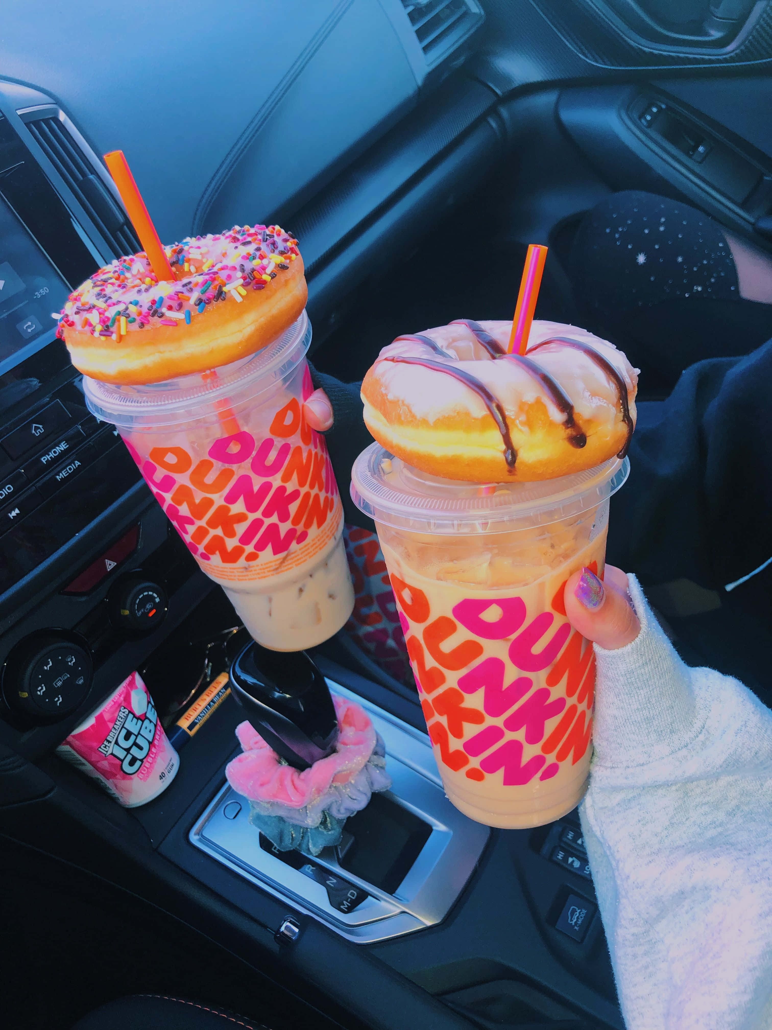 Dunkin Donuts 桌布 壁紙 — 兩隻手在車內拿著兩杯上面有糖霜甜甜圈的Dunkin'冰飲，排檔桿上有一個粉紅色的髮圈。