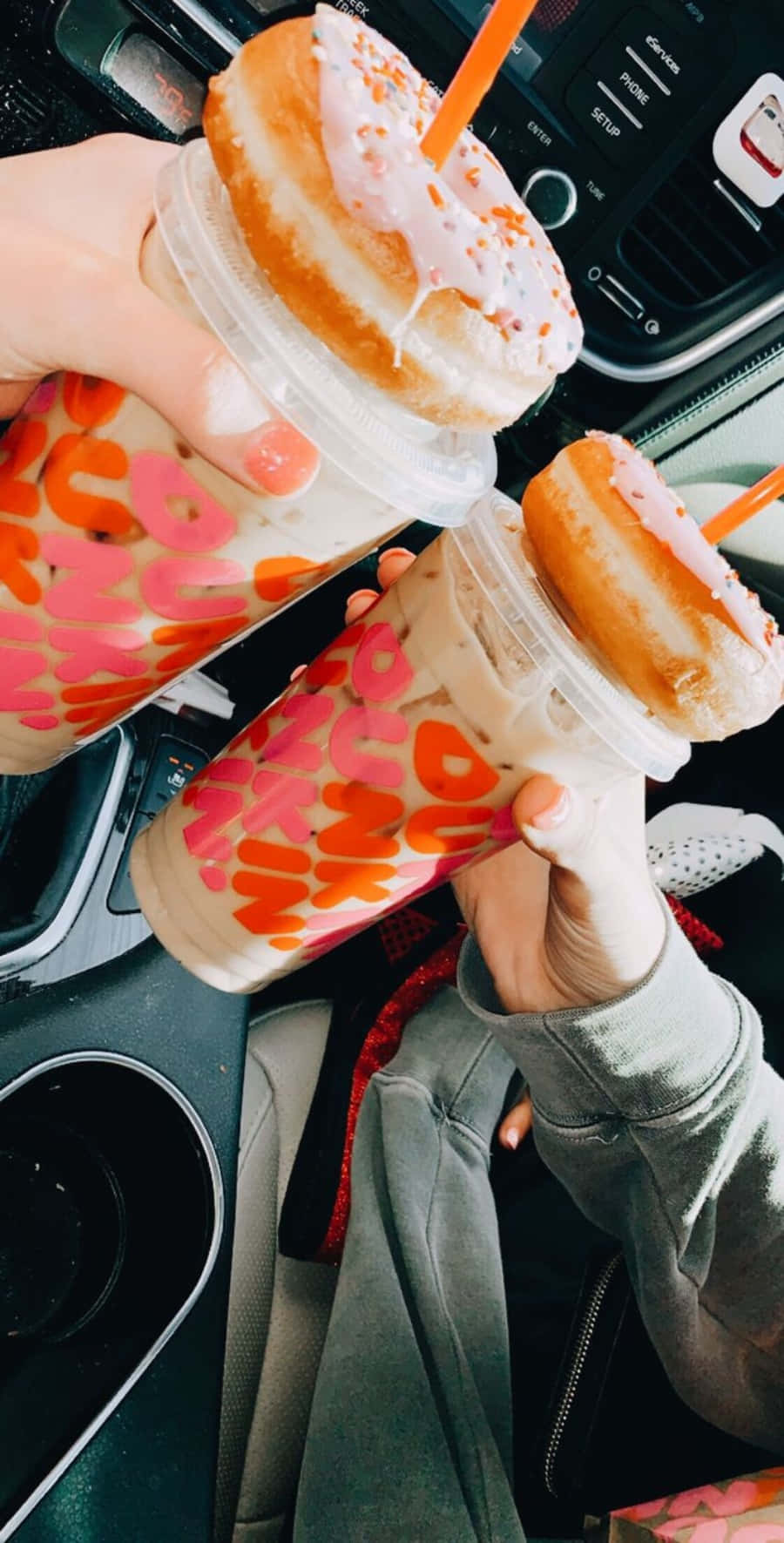 Dunkin Donuts 桌布 壁紙 — 兩隻手在車內拿著相配的冰凍飲料，上面有甜甜圈，車內可見儀表板和杯架。