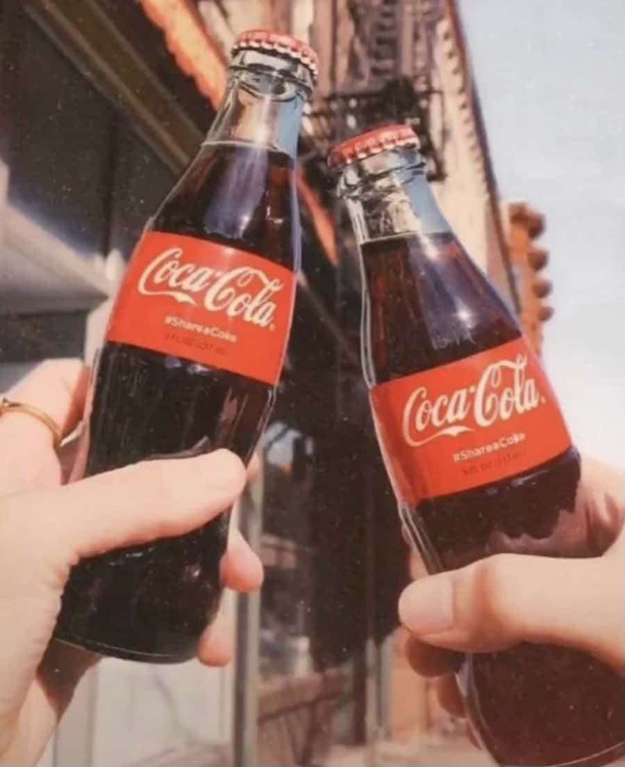 coca cola 桌布 壁紙 — 兩隻手在陽光明媚的城市背景下，碰擊帶有紅色標籤的復古可口可樂玻璃瓶。