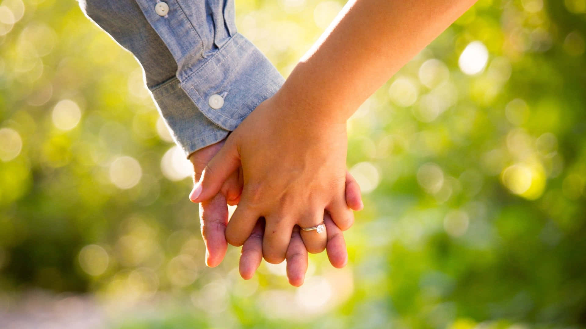 Holding Hands 桌布 壁紙 — 兩隻手在陽光普照的戶外環境中緊握在一起，背景是柔和的綠色散景。