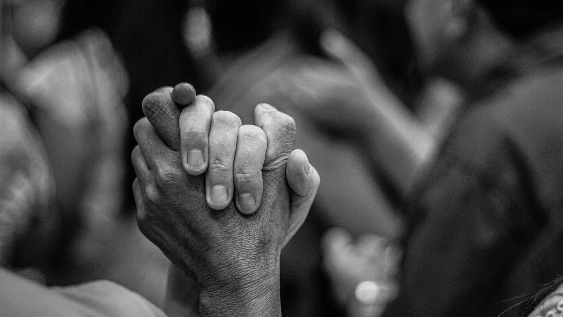 Racism 桌布 壁紙 — 兩隻手在黑白特寫中緊緊相握，在模糊的人群背景前傳達深刻的連結和支持。