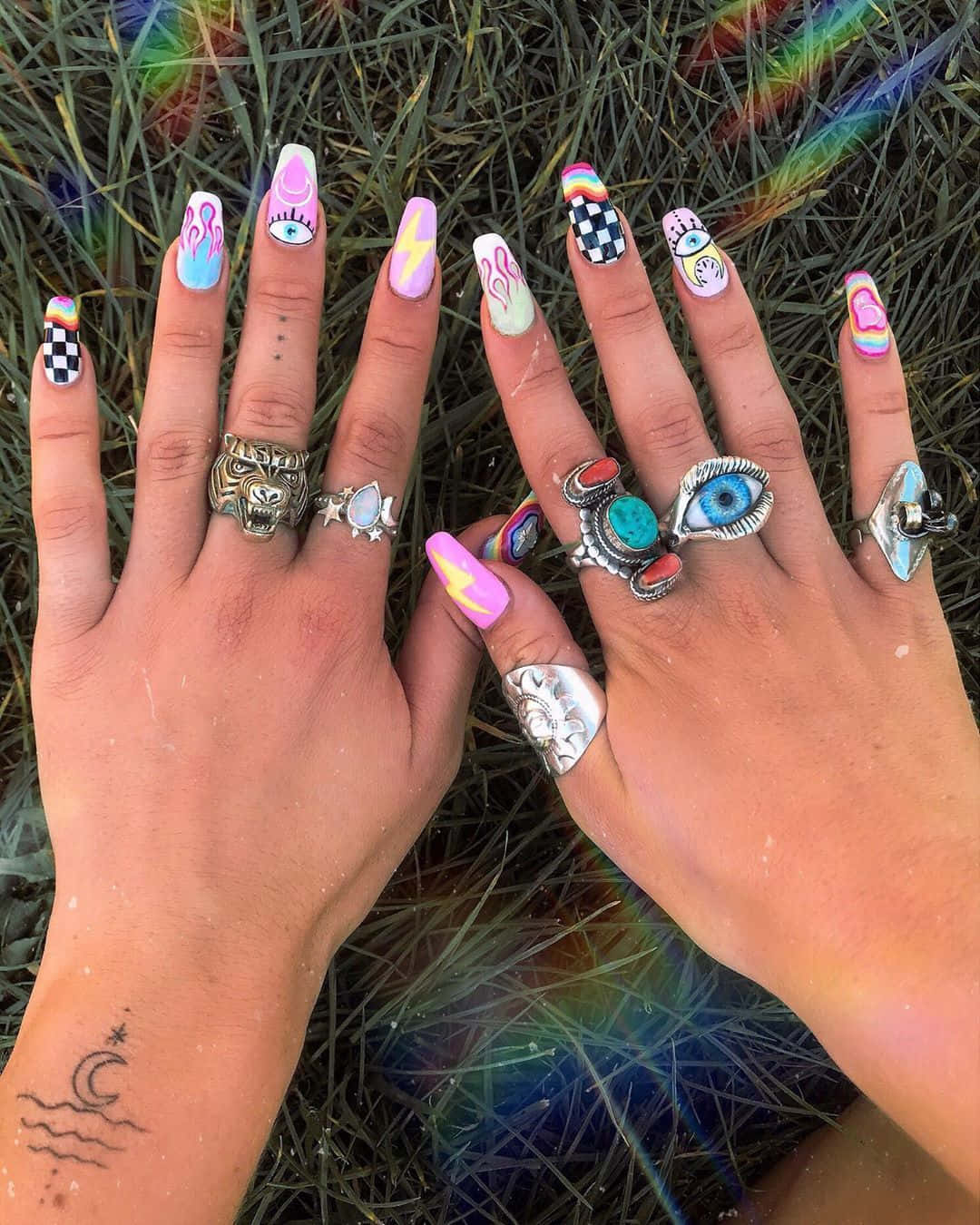 Nail 桌布 壁紙 — 兩隻手帶有色彩繽紛的迷幻指甲藝術和波希米亞戒指，放在草地上，配以彩虹羽毛點綴。
